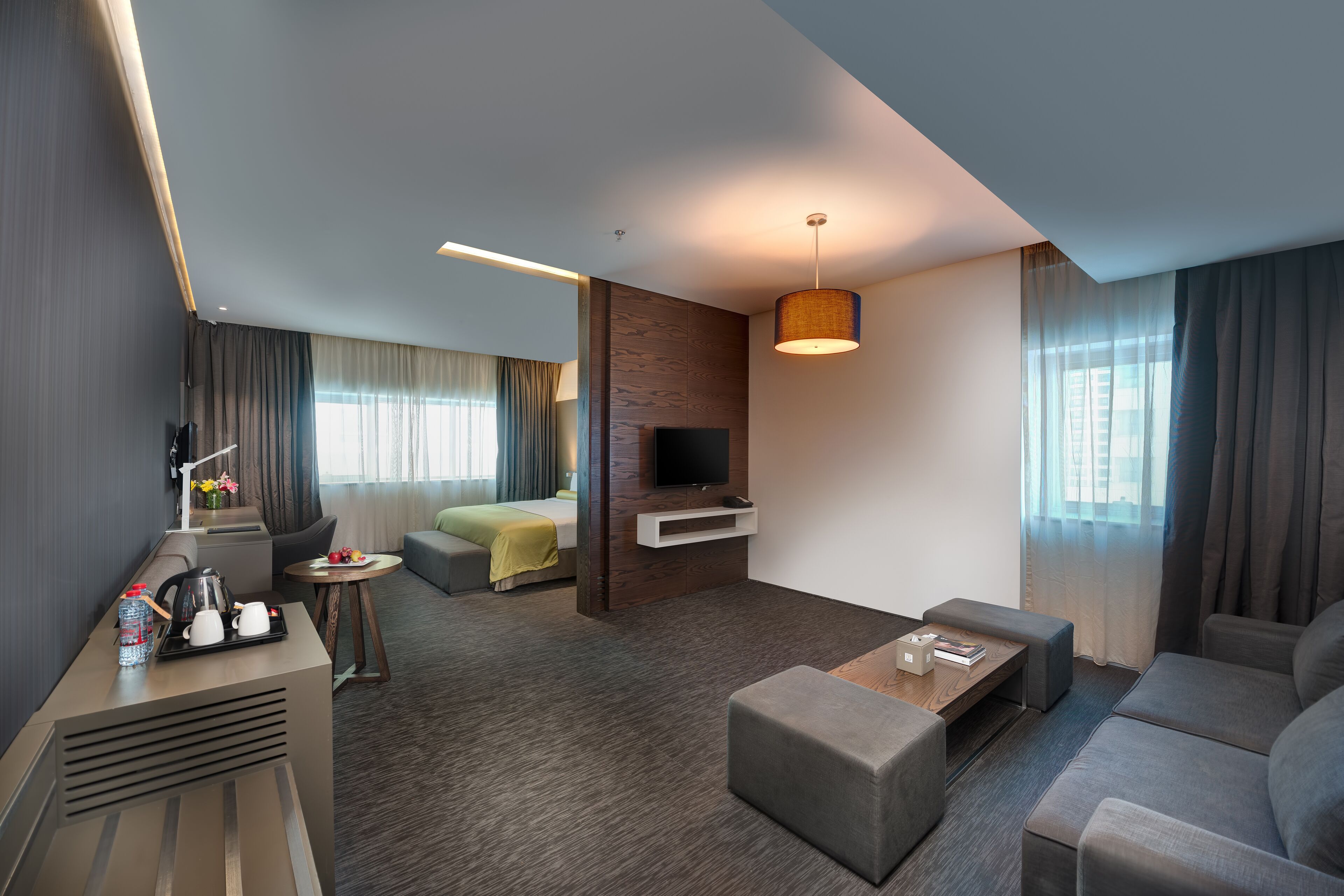 Quarto de hotel moderno com sofá, mesa de centro, TV fixada na parede, pequena mesa lateral com abajur e cama com manta verde.