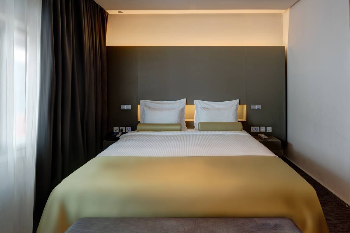 A hotel room with a large bed, two bedside tables, a nightstand, and a window with curtains.