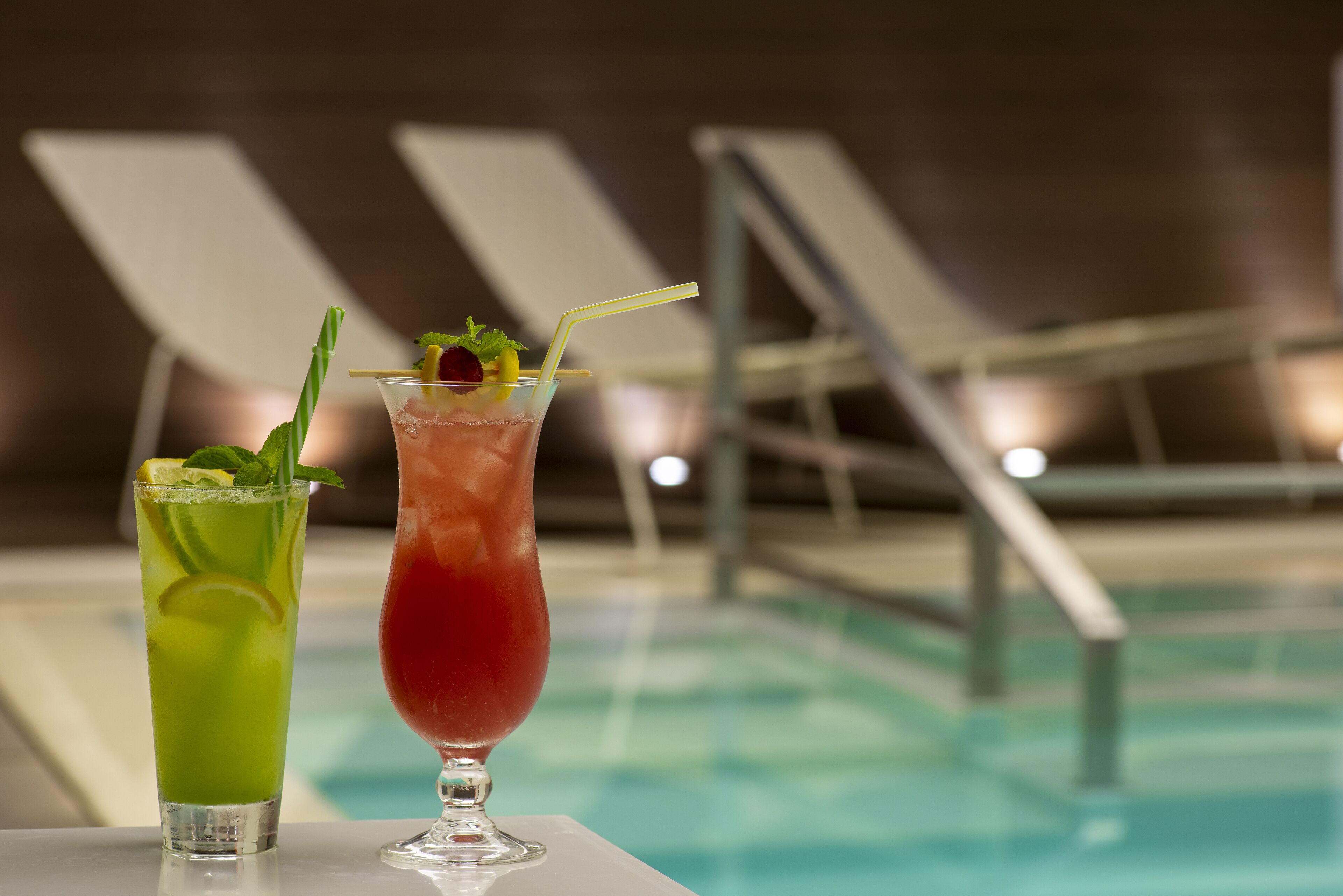 Two cocktails on a table by a poolside with lounge chairs in the background.