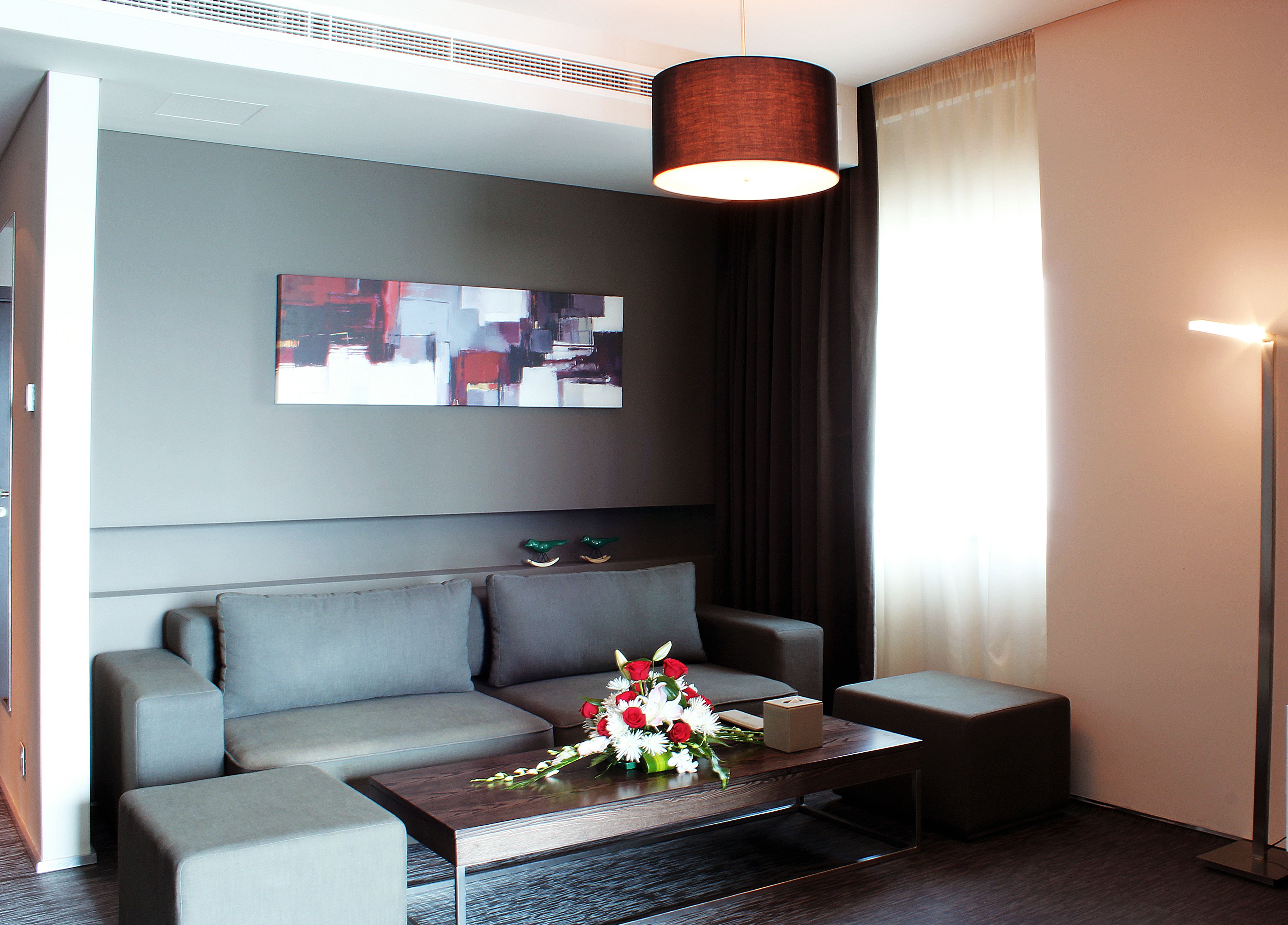 A modern hotel room with a grey sofa, a wooden coffee table with a floral arrangement, and a large abstract painting on the wall.