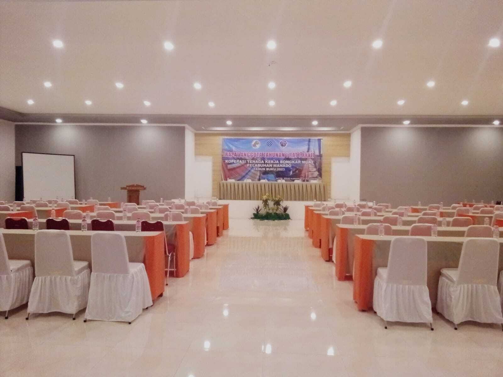 A conference room with rows of tables and chairs, a podium, and a large screen at the front.