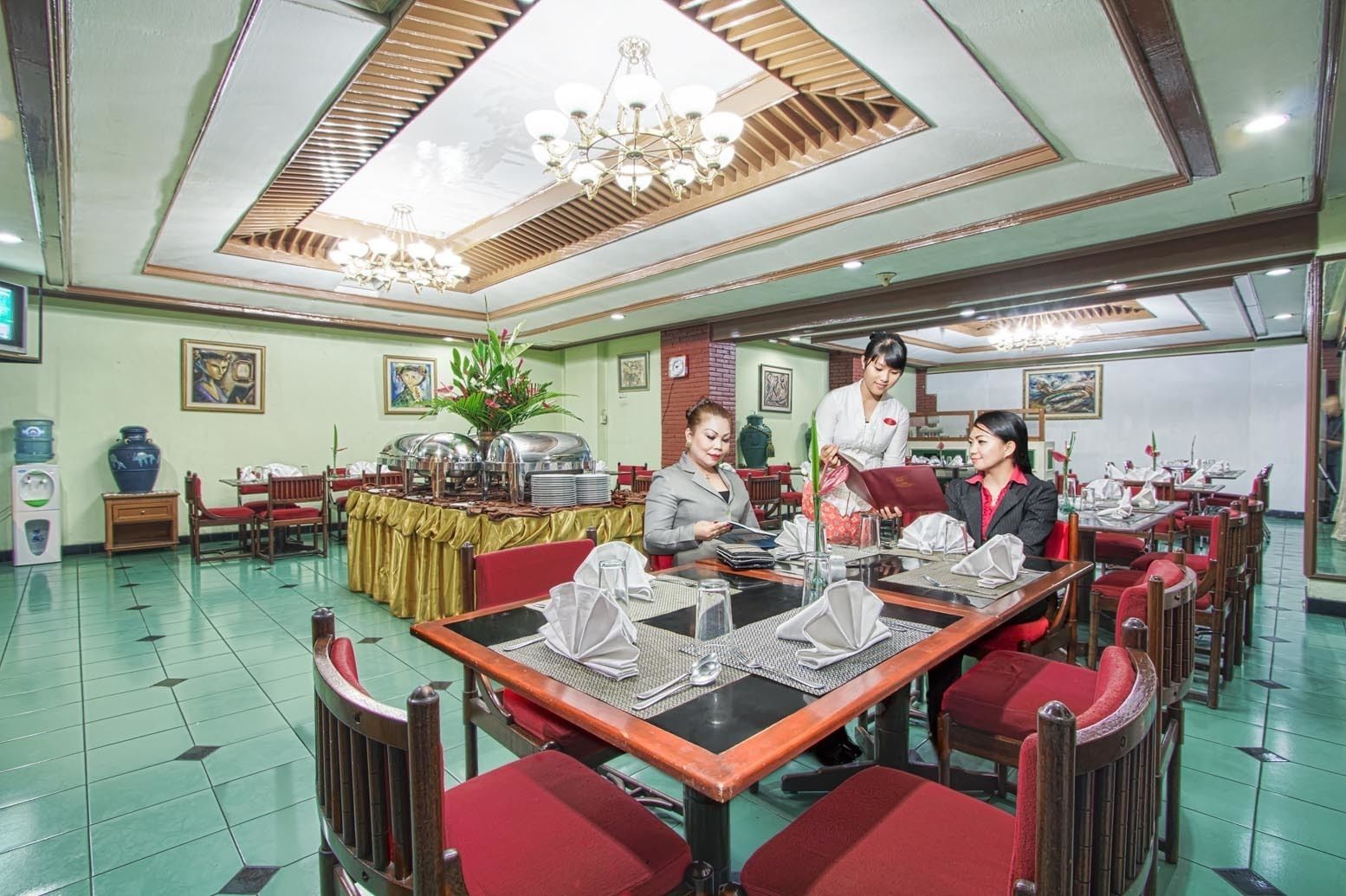 Comedor con mesas dispuestas para el servicio, un bufé con recipientes de comida y un miembro del personal atendiendo a los huéspedes.