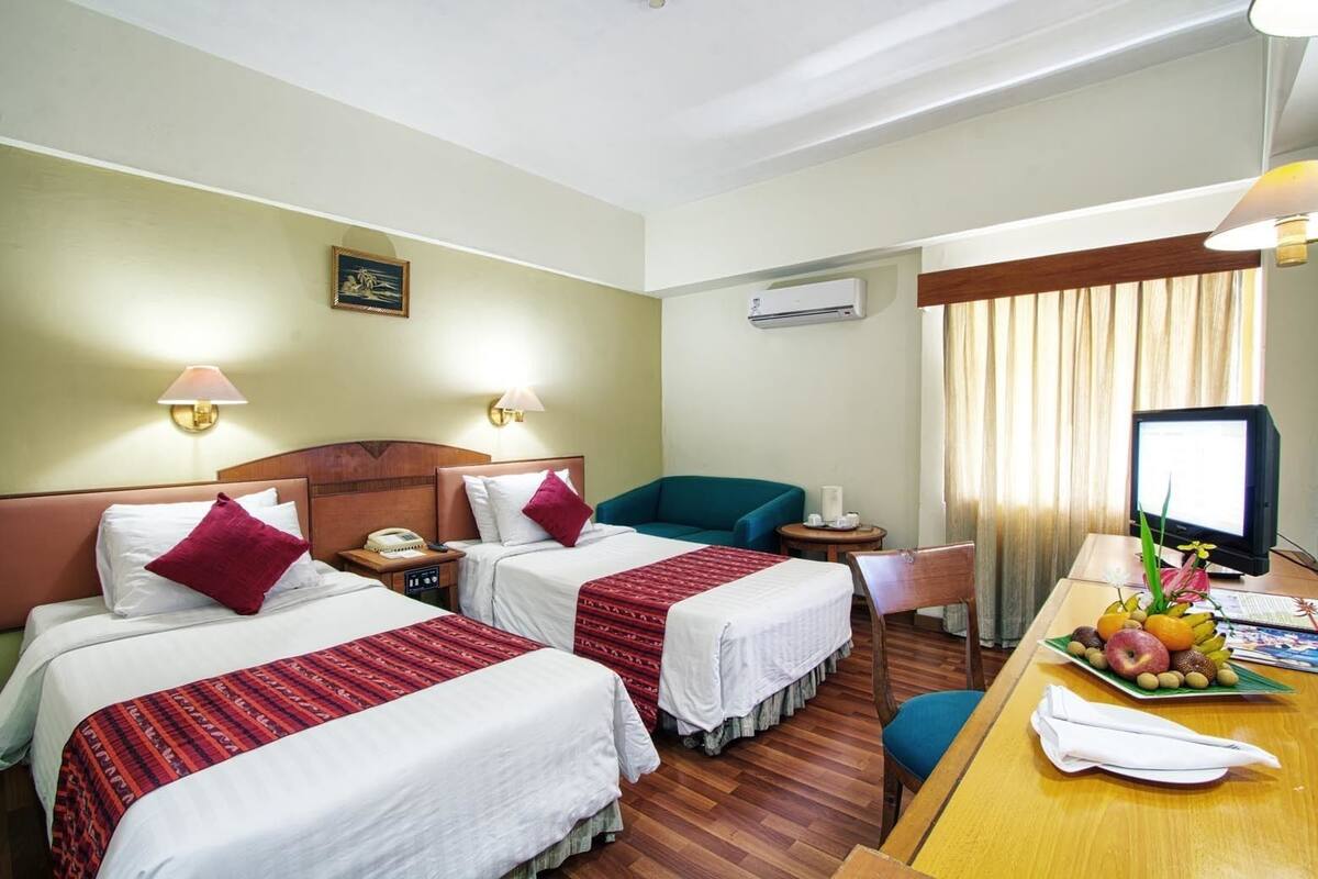 A hotel room with two beds, a desk with a fruit bowl, a television, and an air conditioning unit.