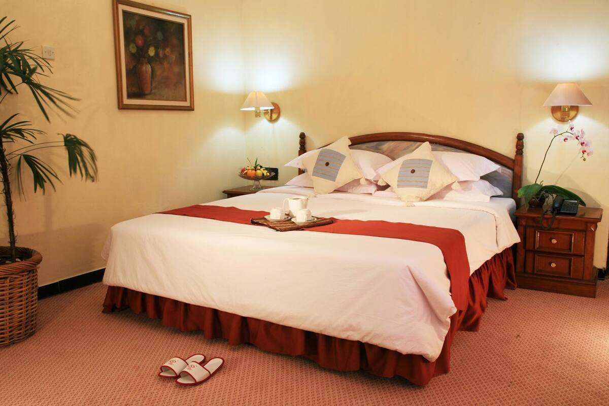 A neatly made bed with a red skirt, a bedside table with a lamp, a fruit bowl, and a potted plant.