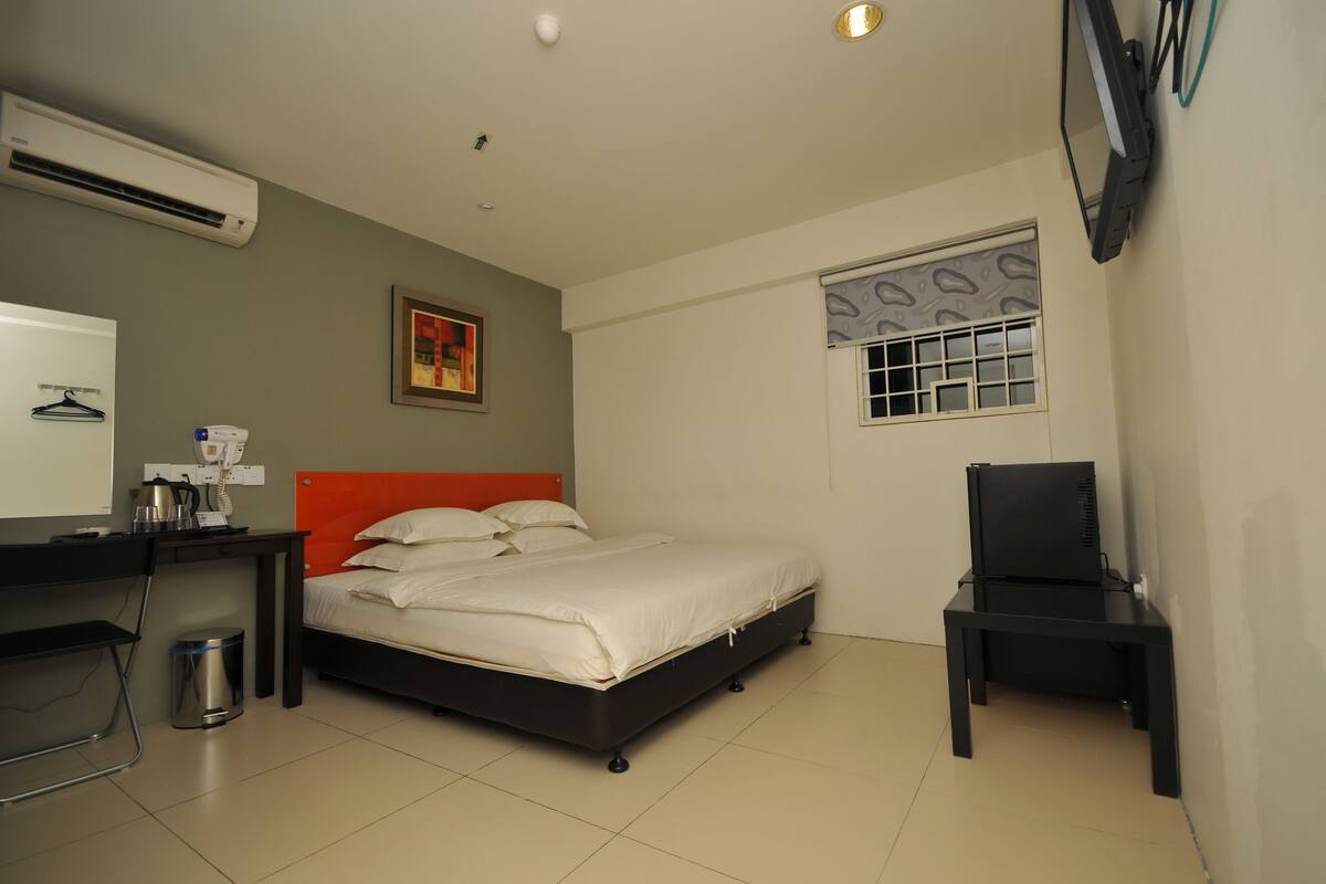 Quarto de hotel com cama, escrivaninha, cadeira, ar condicionado, lixeira e janela com persiana estampada.