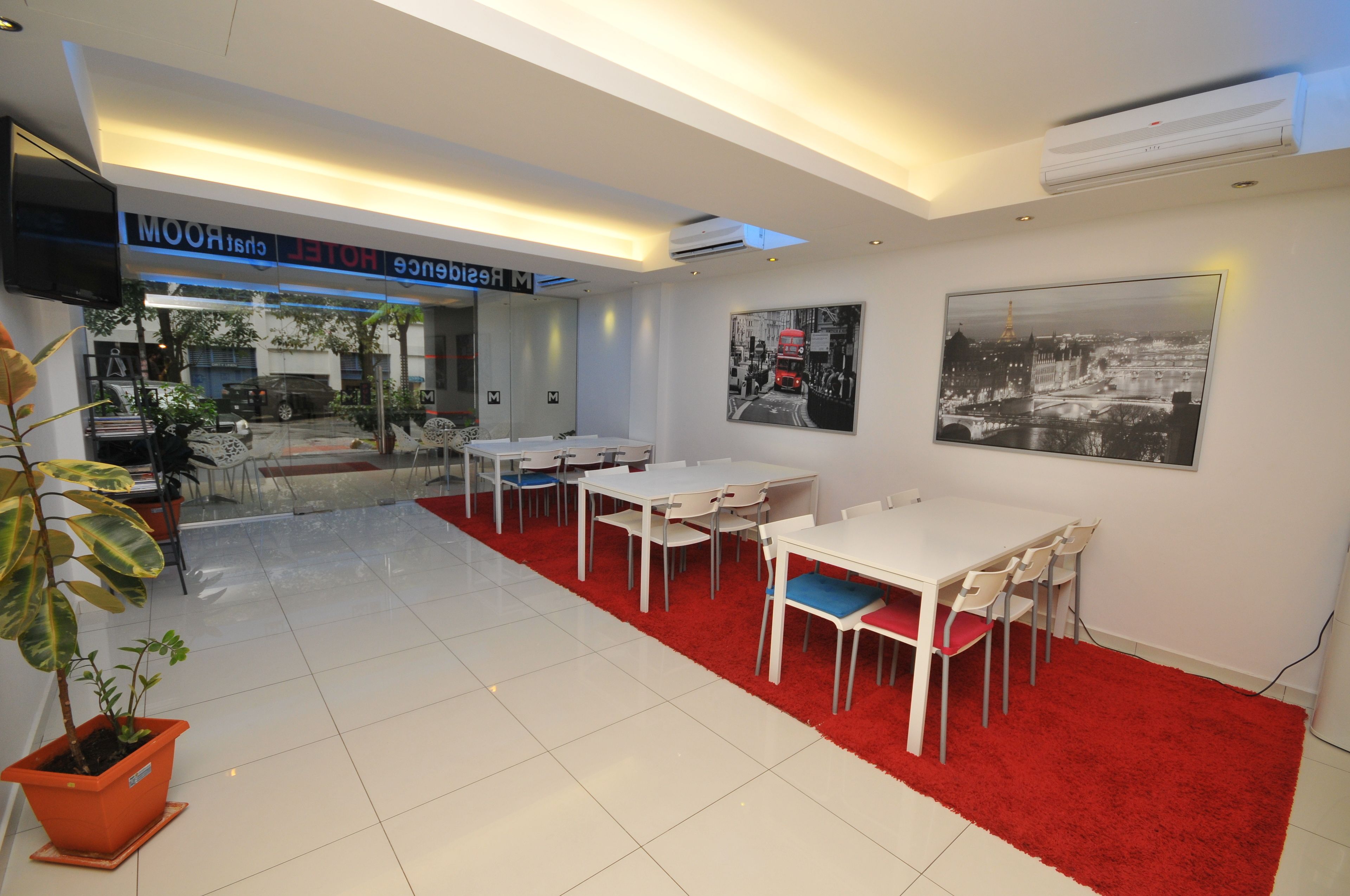 Een hotellobby met een rood tapijt, witte tafels en stoelen, een groot zwart-wit stadsgezicht schilderij en een airconditioningsunit aan het plafond.