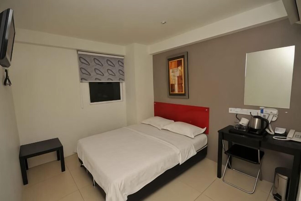 A hotel room with a bed, a desk, a chair, a small table, a television, and a window with blinds.