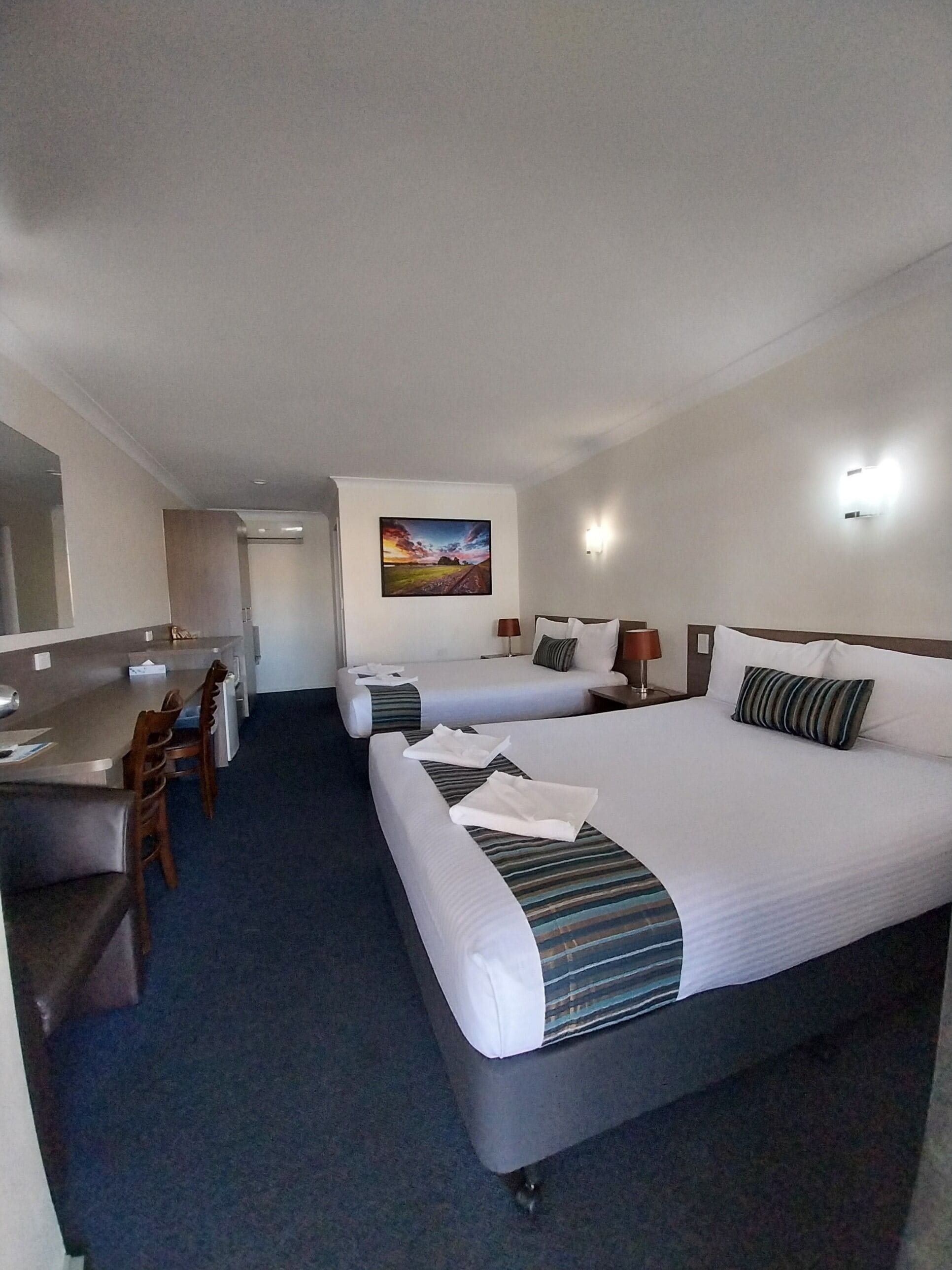 A hotel room with two beds, a desk, and a chair. There is a painting on the wall and a lamp on the desk.