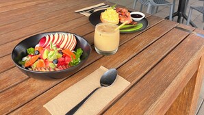 Ein Holztisch mit Salat, einem Getränk und einer Speisenplatte. Im Hintergrund sind Blumentöpfe und eine Glastür zu sehen.