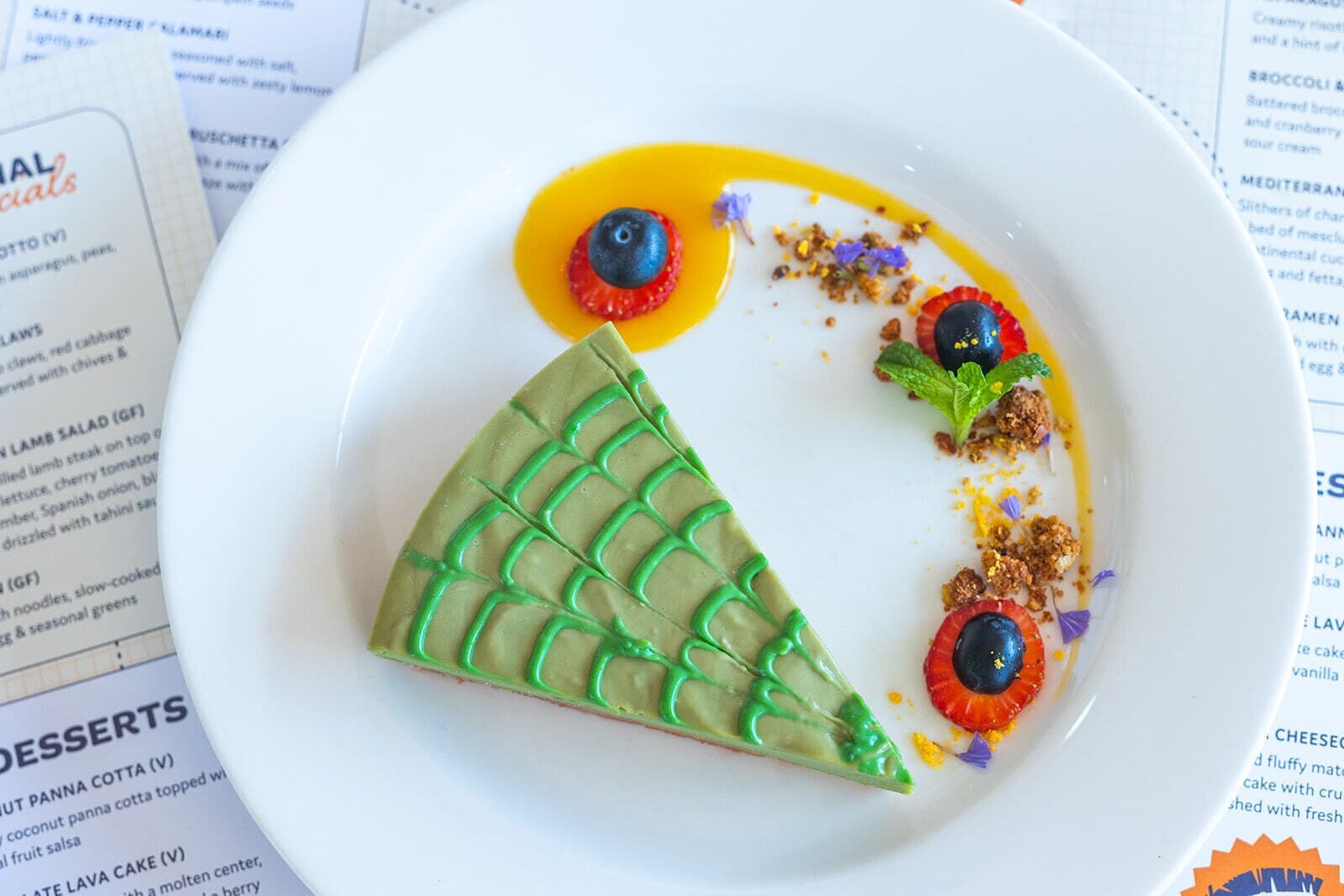 A dessert plate with a green triangular pastry, blueberries, and a yellow sauce.