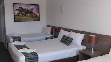 A hotel room with three beds, a painting on the wall, and a lamp on the nightstand.