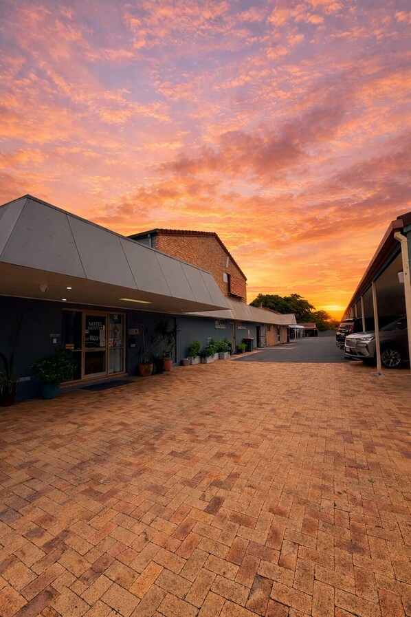 A motel with a modern design, featuring a brick-paved driveway and a clear view of the sunset in the background.