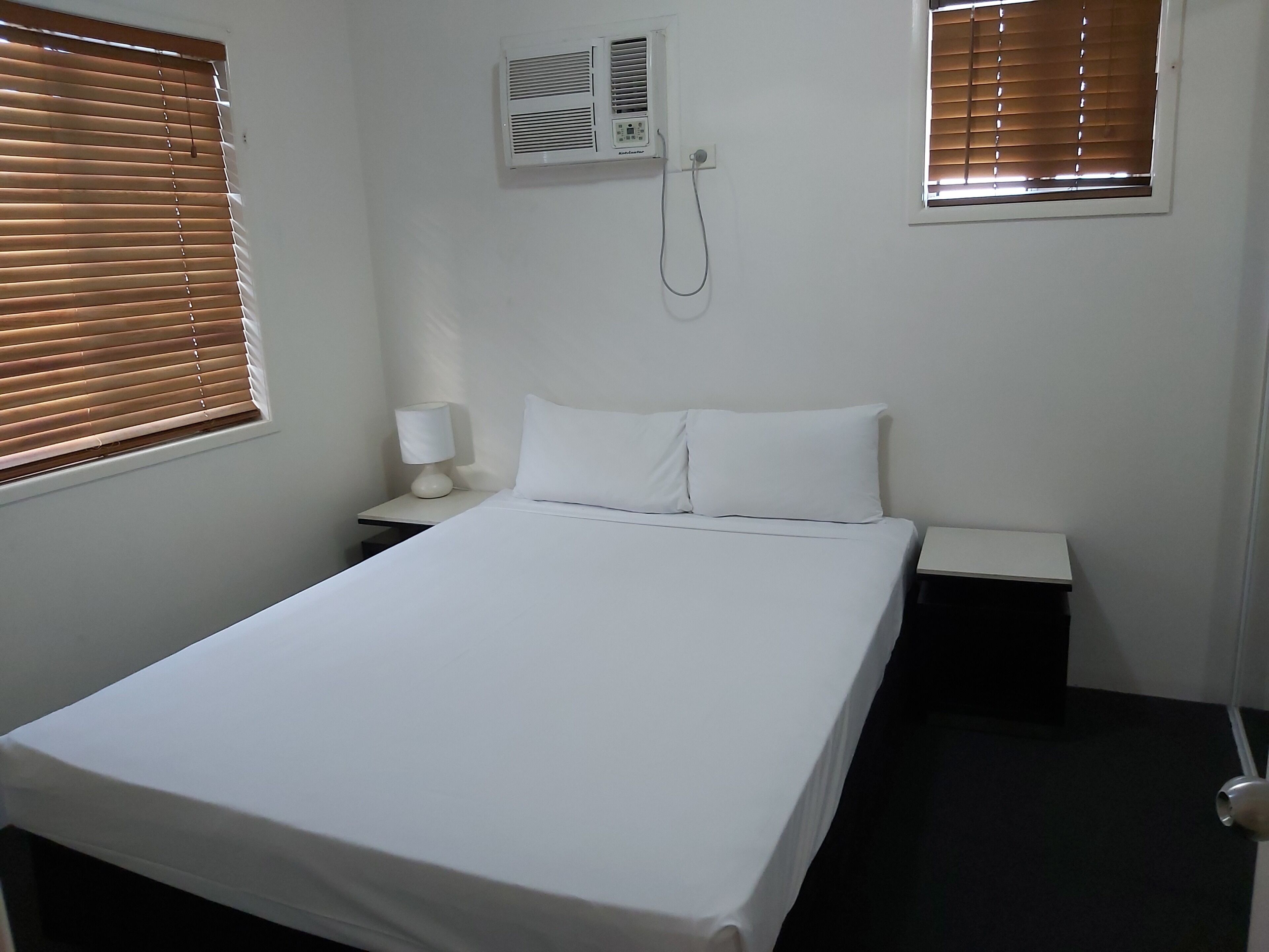 Una habitación de hotel con una cama individual, una mesita de noche con lámpara, un aire acondicionado instalado en la pared y dos ventanas con persianas.