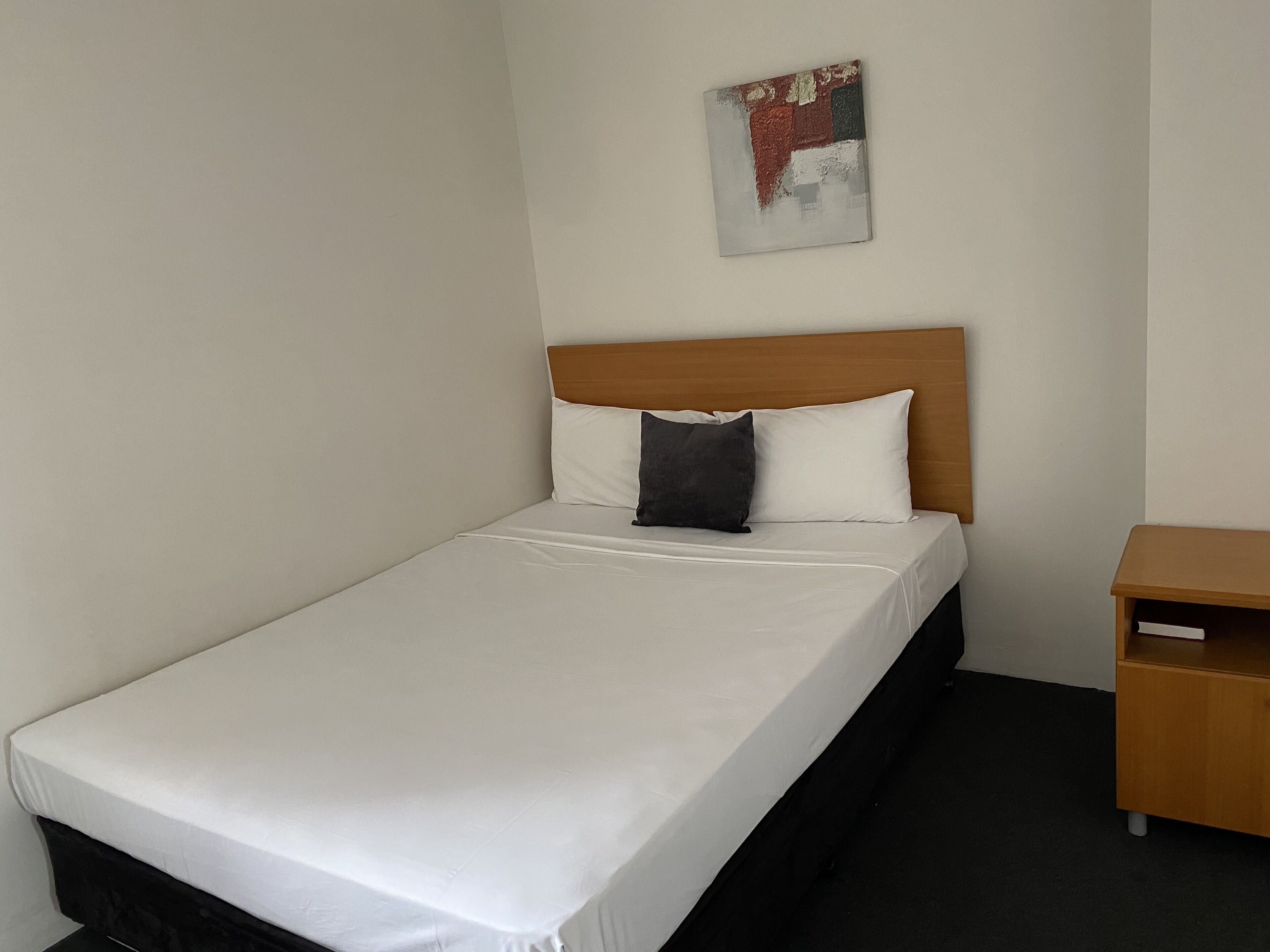 A single bed with a wooden headboard, white bedding, and a small wooden nightstand with a drawer.
