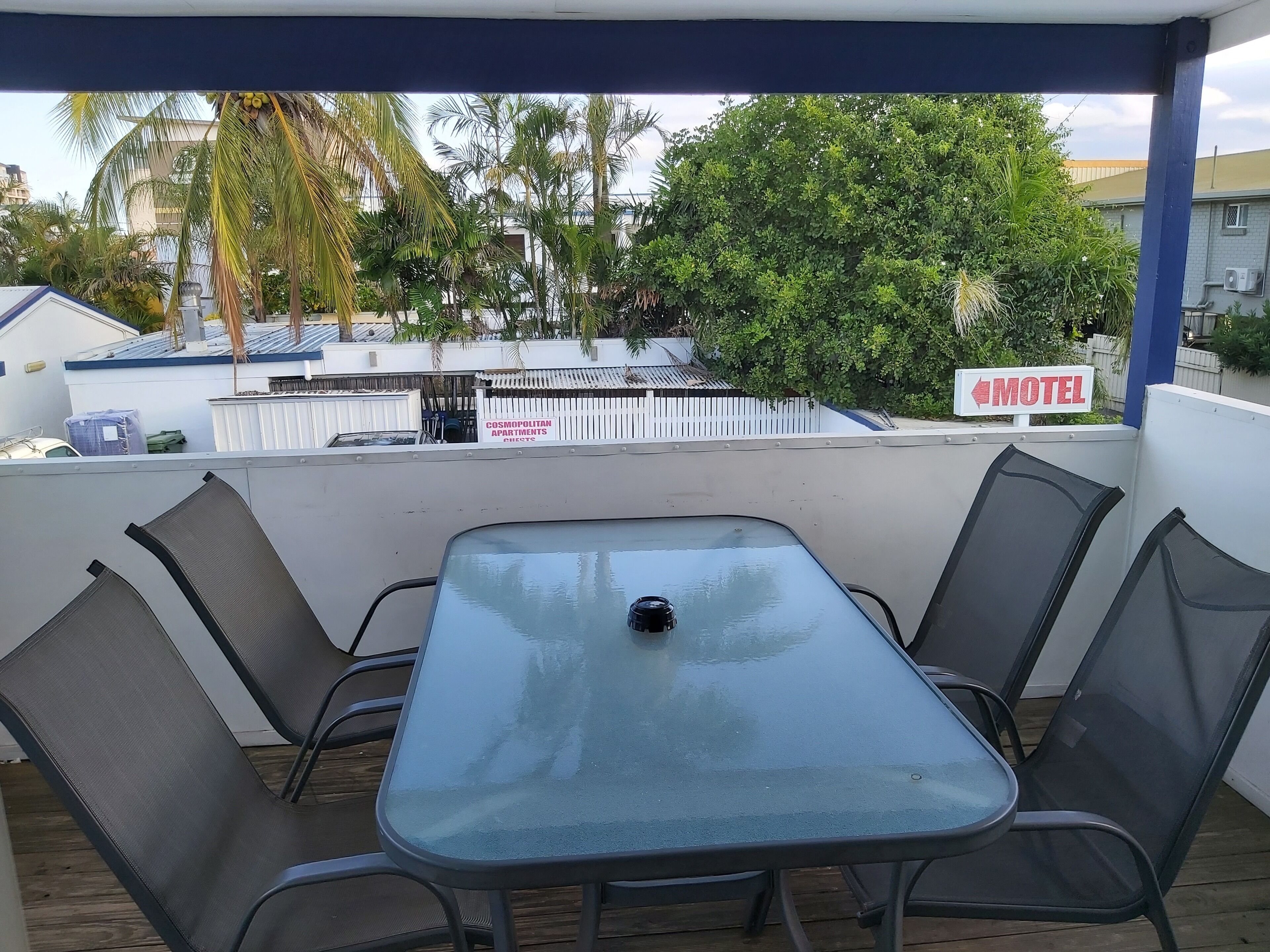 A covered outdoor seating area with a table and chairs, overlooking a fenced area with a 'Motel' sign and 'Cosmopolitan Apartments' in the background.