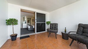 A modern living room with a sliding glass door leading to a bedroom, a grey armchair, a wicker chair, and two potted plants. - The Cosmopolitan Motel and Serviced Apartments (Rockhampton)