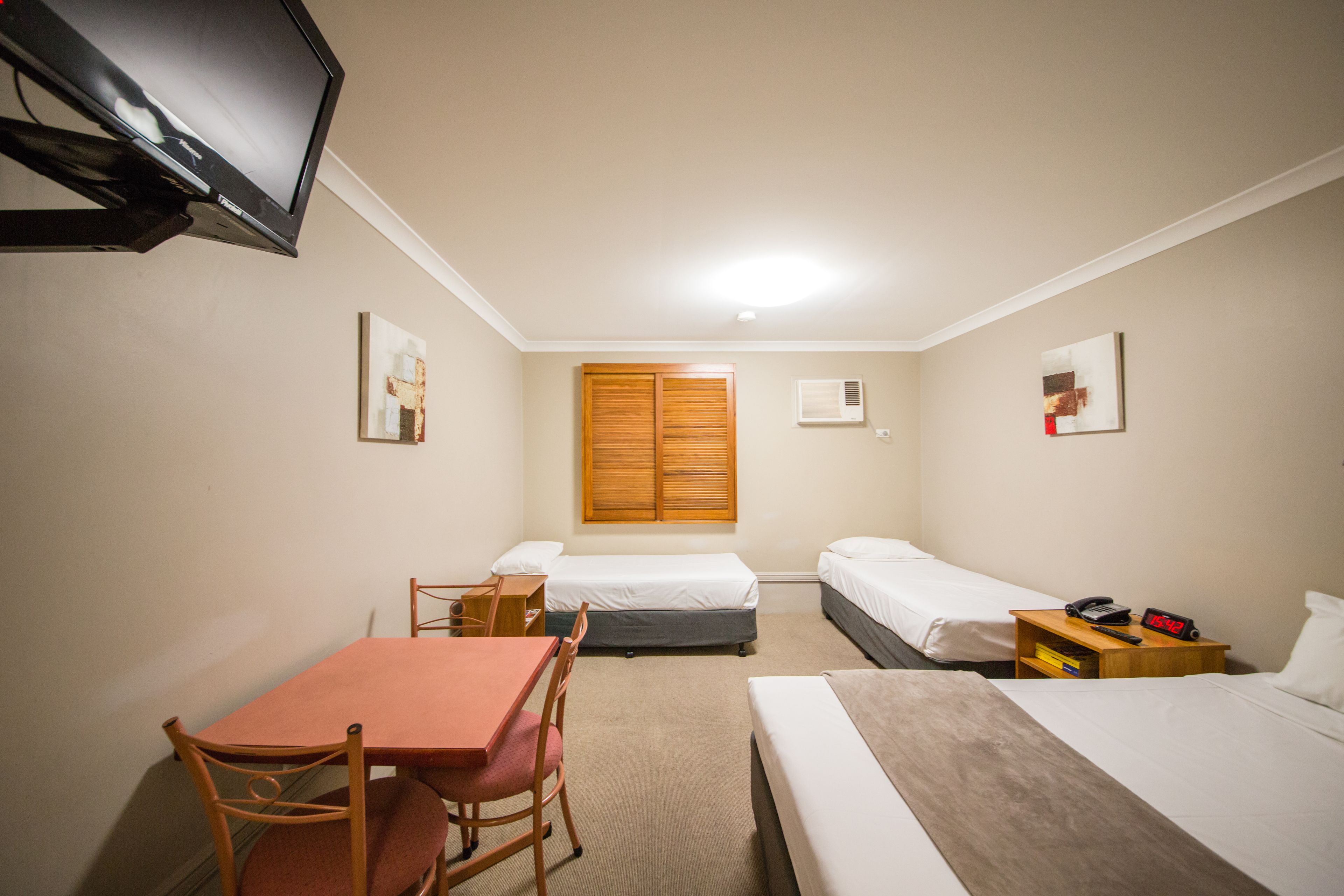 Une chambre d’hôtel avec un lit simple, un lit double, un bureau avec une chaise, un téléviseur fixé au mur et une armoire en bois.