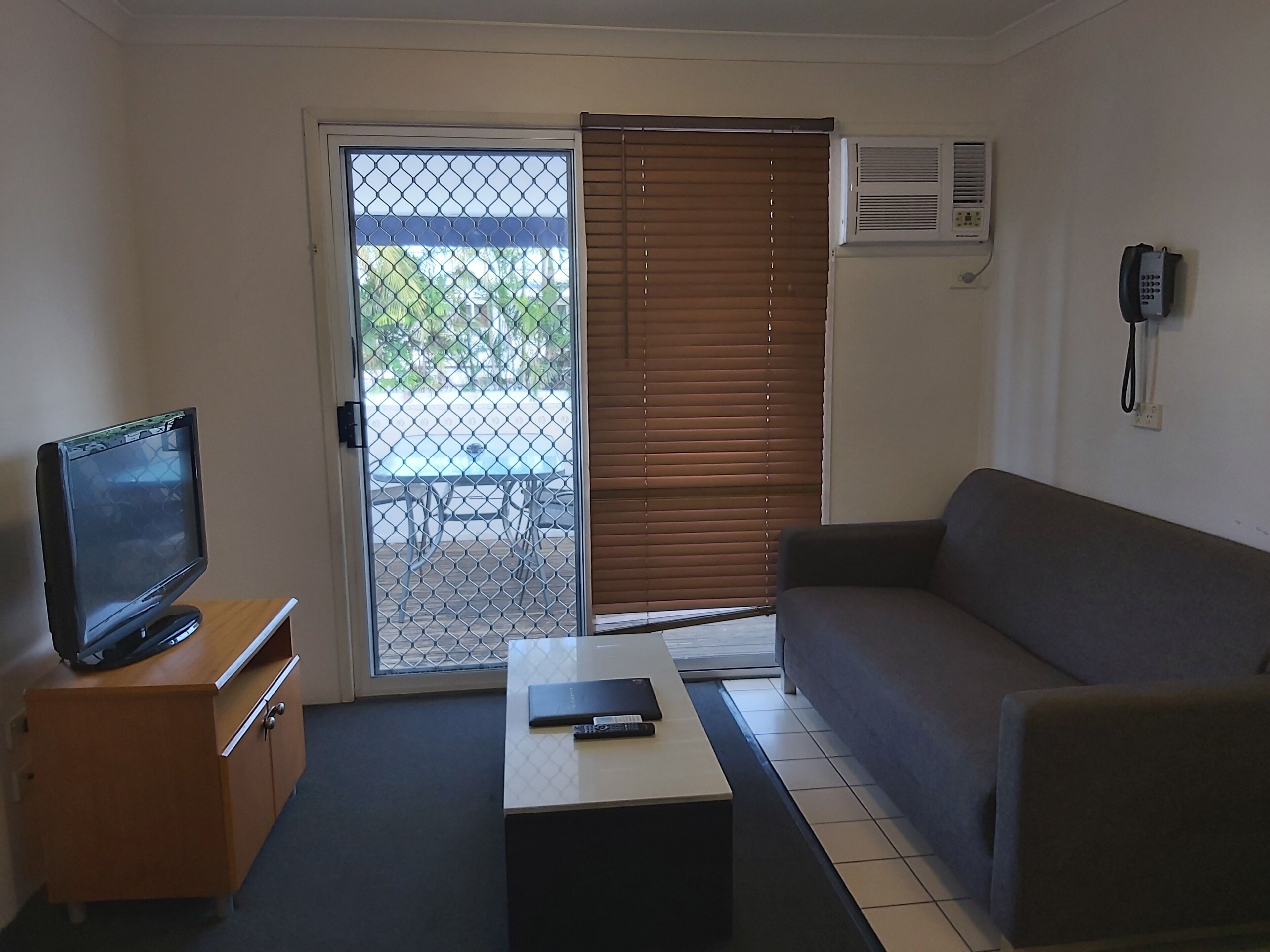 A room with a television on a wooden cabinet, a sofa, a coffee table, and a sliding glass door leading to an outdoor area with a table and chairs.