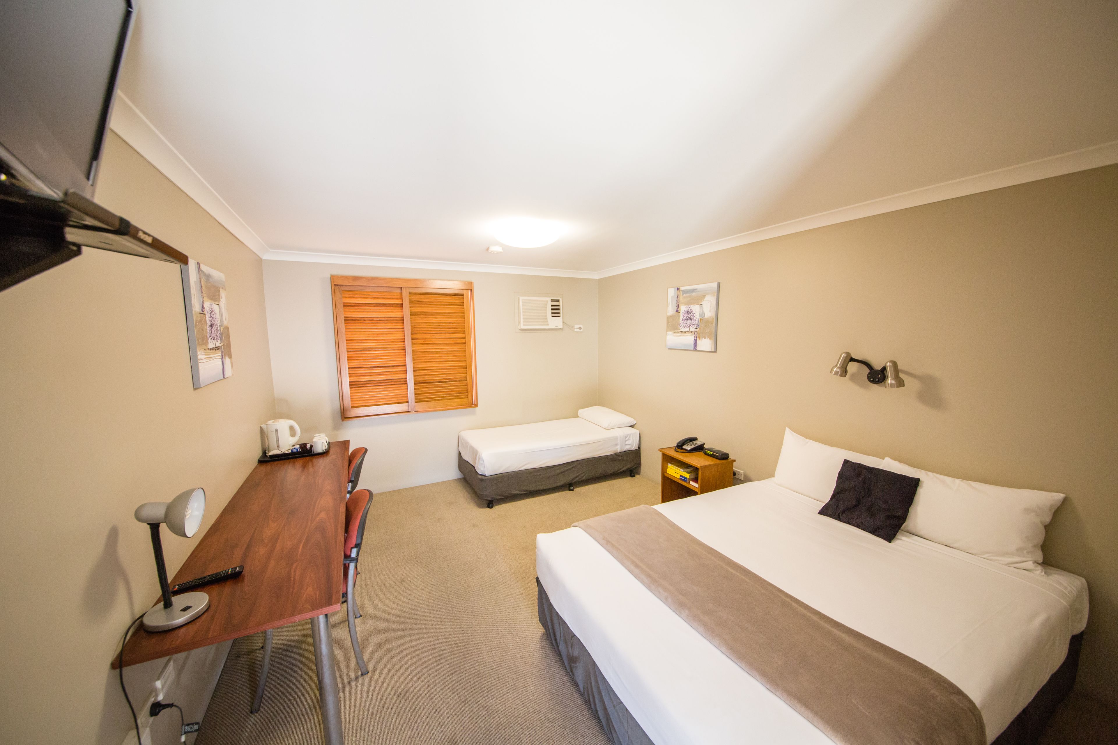 A hotel room with a bed, a desk with a chair, a small bedside table with a phone, a window with blinds, and a wall-mounted light fixture.