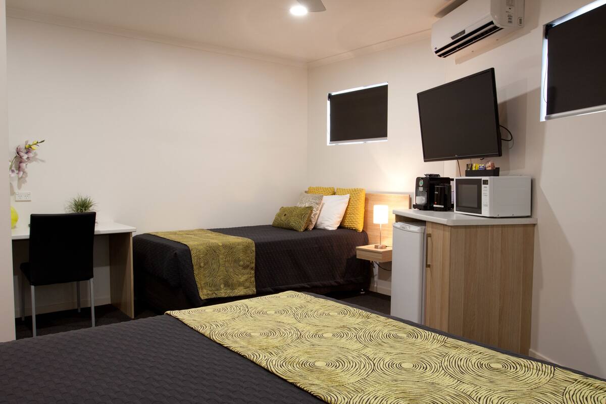 A compact hotel room with a bed, a desk, a chair, a small kitchenette, and a TV mounted on the wall.