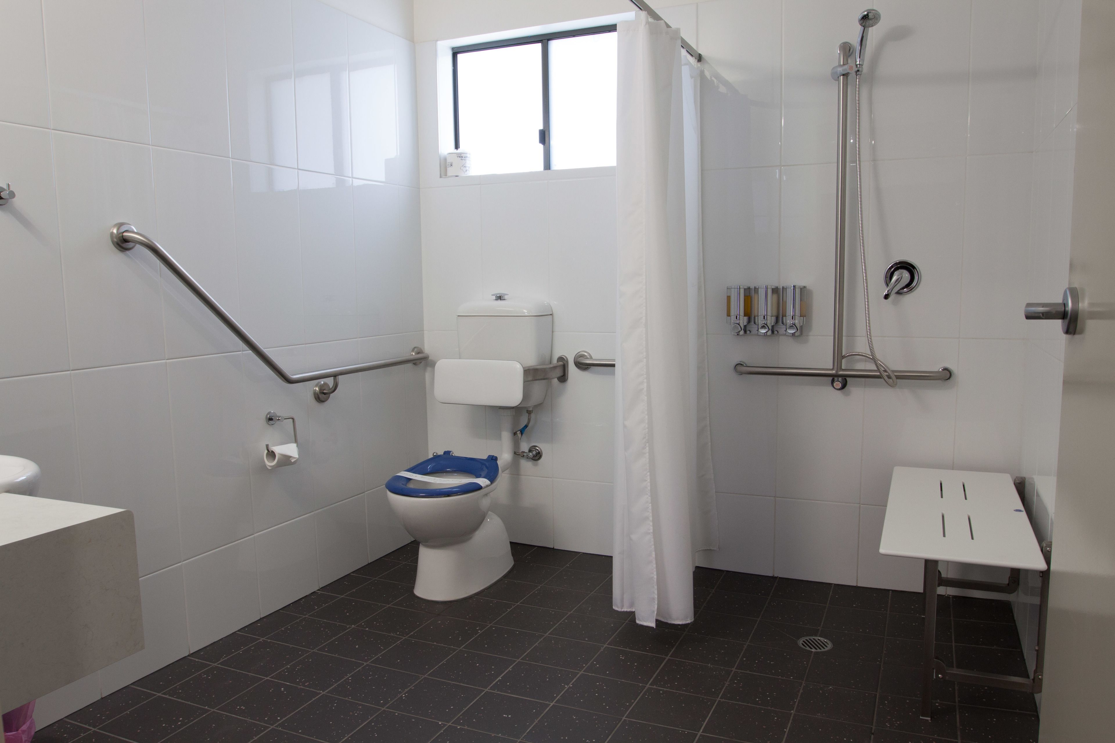 A bathroom with a toilet, a shower, a grab bar, a window, and a small table.