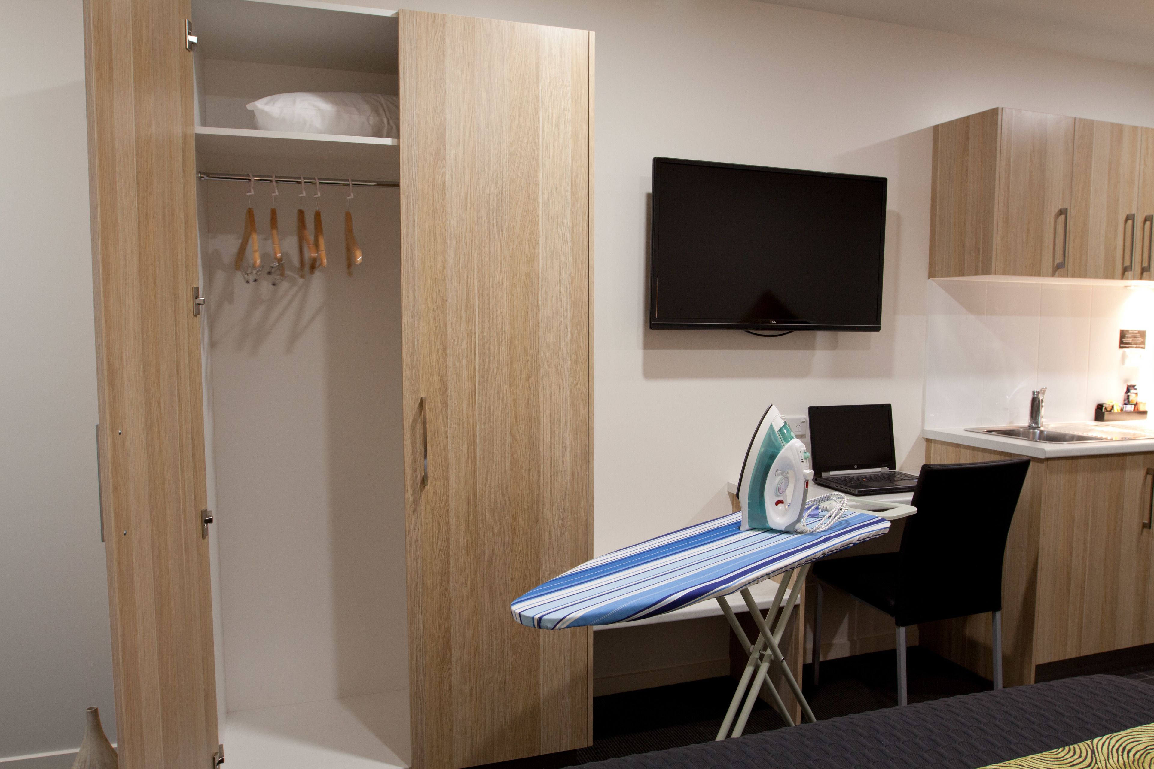 A compact kitchen with a flat-screen TV, a laptop, and an ironing station.