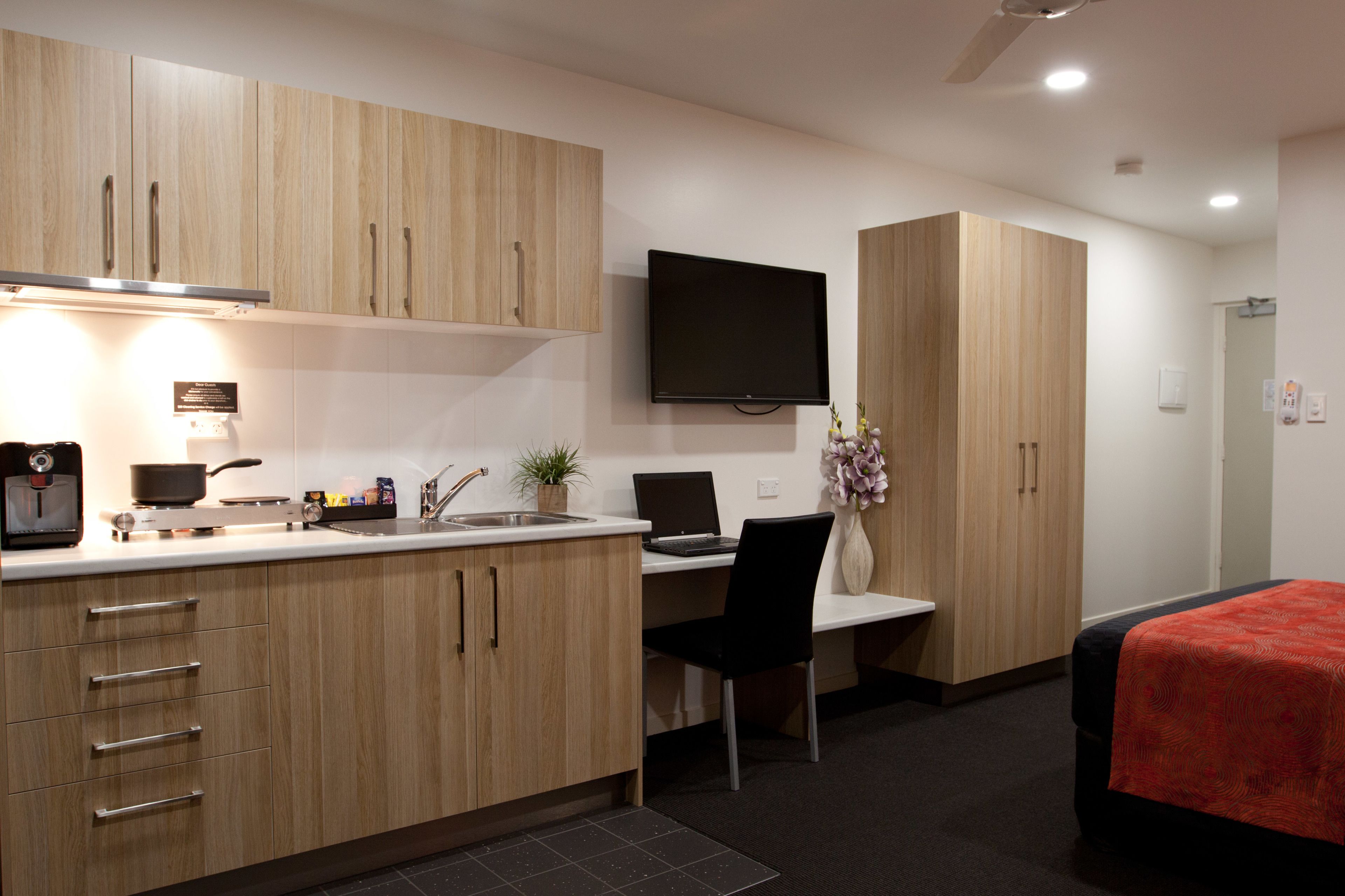 A modern hotel room with a kitchenette, a flat-screen TV, a bed with a red blanket, and a desk with a laptop.