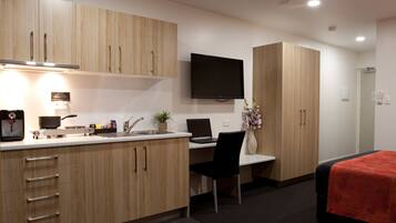 A modern hotel room with a kitchenette, a flat-screen TV, a bed with a red blanket, and a desk with a laptop.