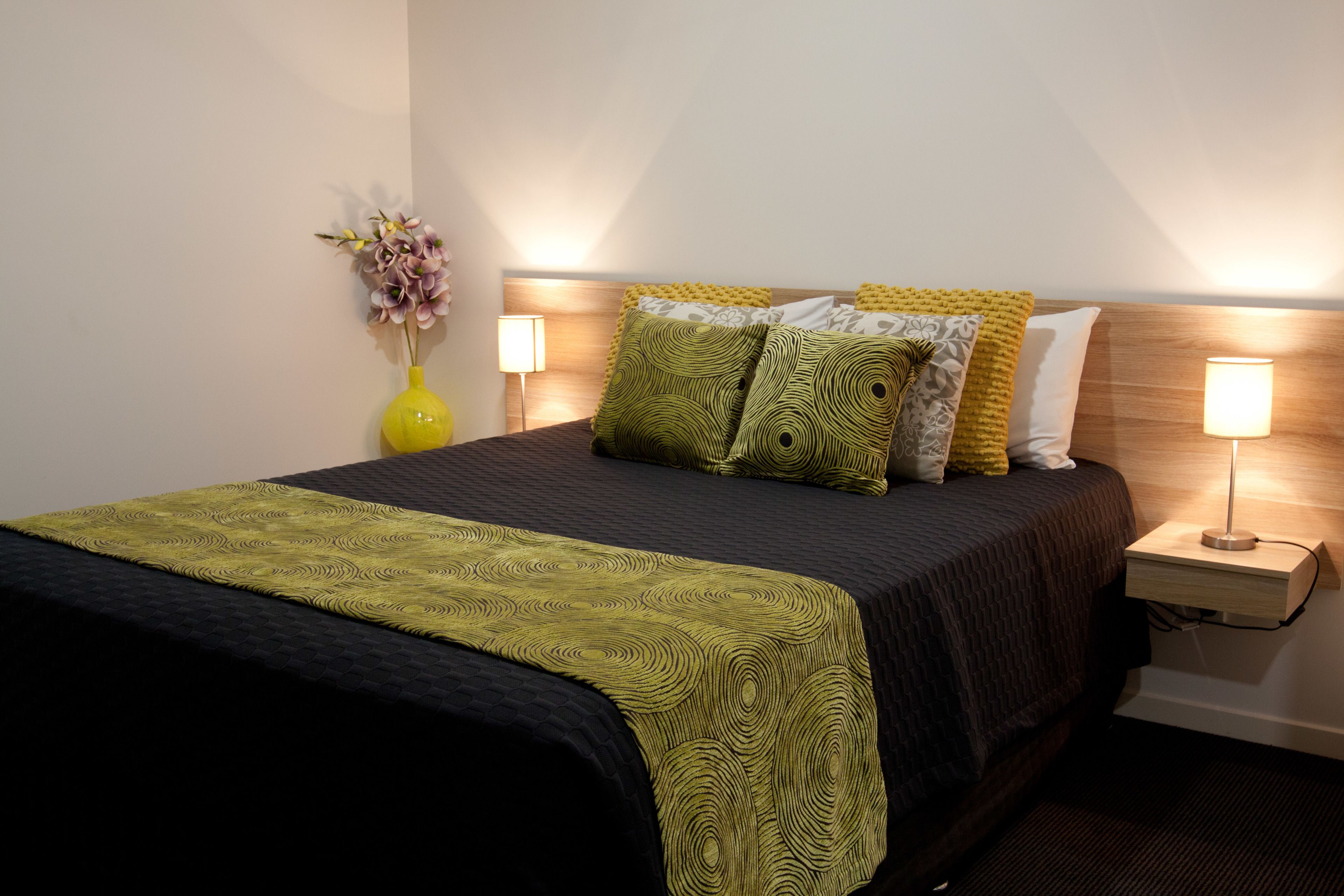 A neatly made bed with a patterned bedspread and pillows, a bedside table with a lamp, and a vase with flowers on the nightstand.