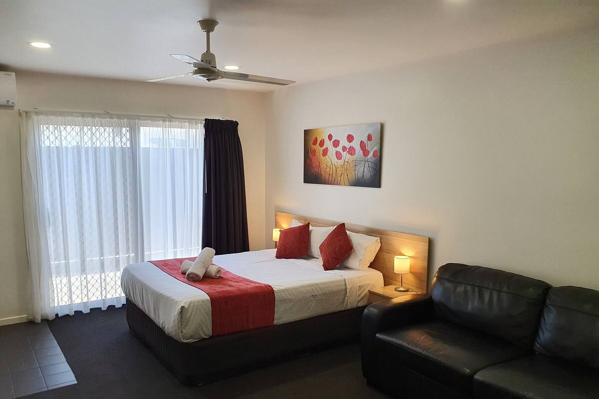A hotel room with a bed, a black leather sofa, a ceiling fan, a painting on the wall, and a window with curtains.