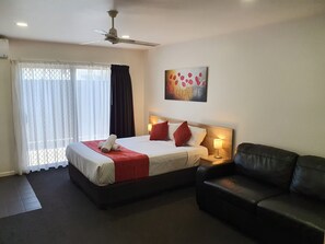 A hotel room with a bed, a black leather sofa, a ceiling fan, a painting on the wall, and a window with curtains.