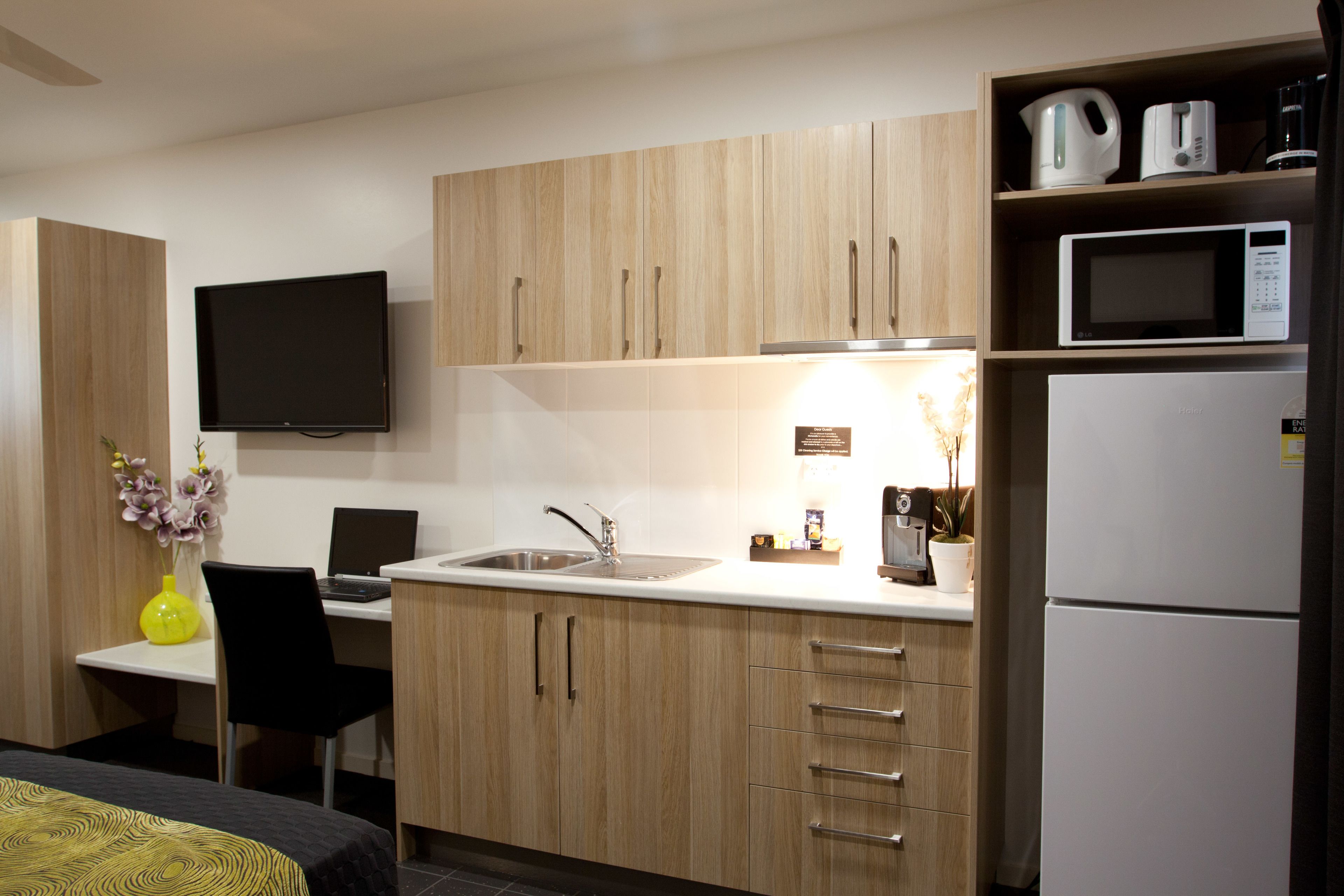 A compact kitchen with wooden cabinets, a microwave, a refrigerator, a sink, and a flat-screen TV.