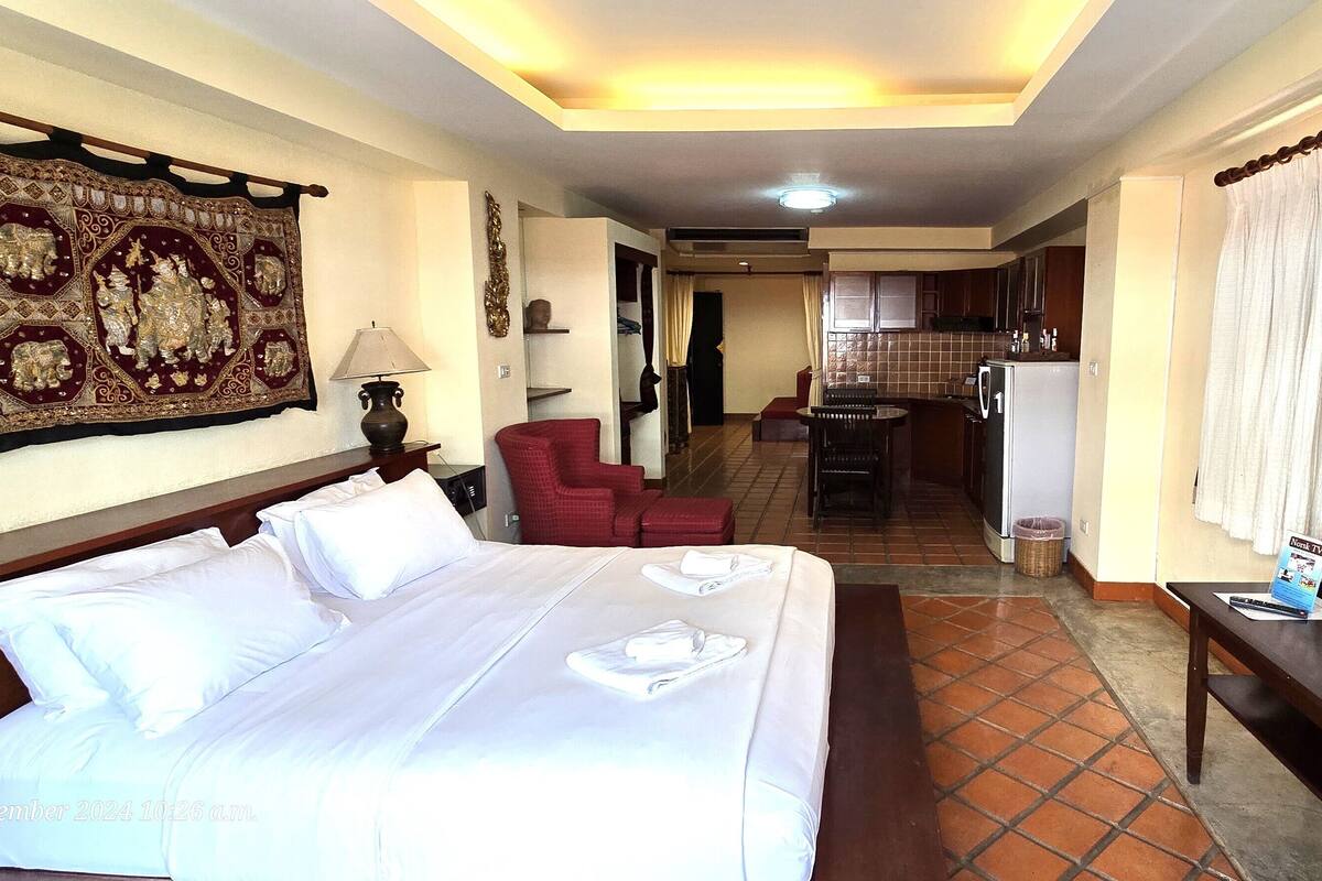 A hotel room with a large bed, a desk, a chair, a TV, and a kitchen area in the background.
