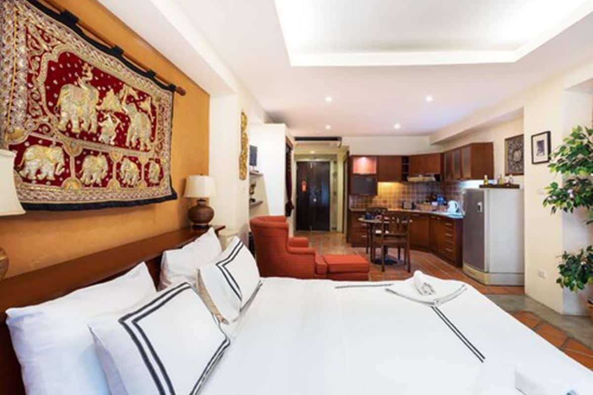 A hotel room with a large bed, a desk, a chair, a red sofa, a kitchen area, and a dining table.