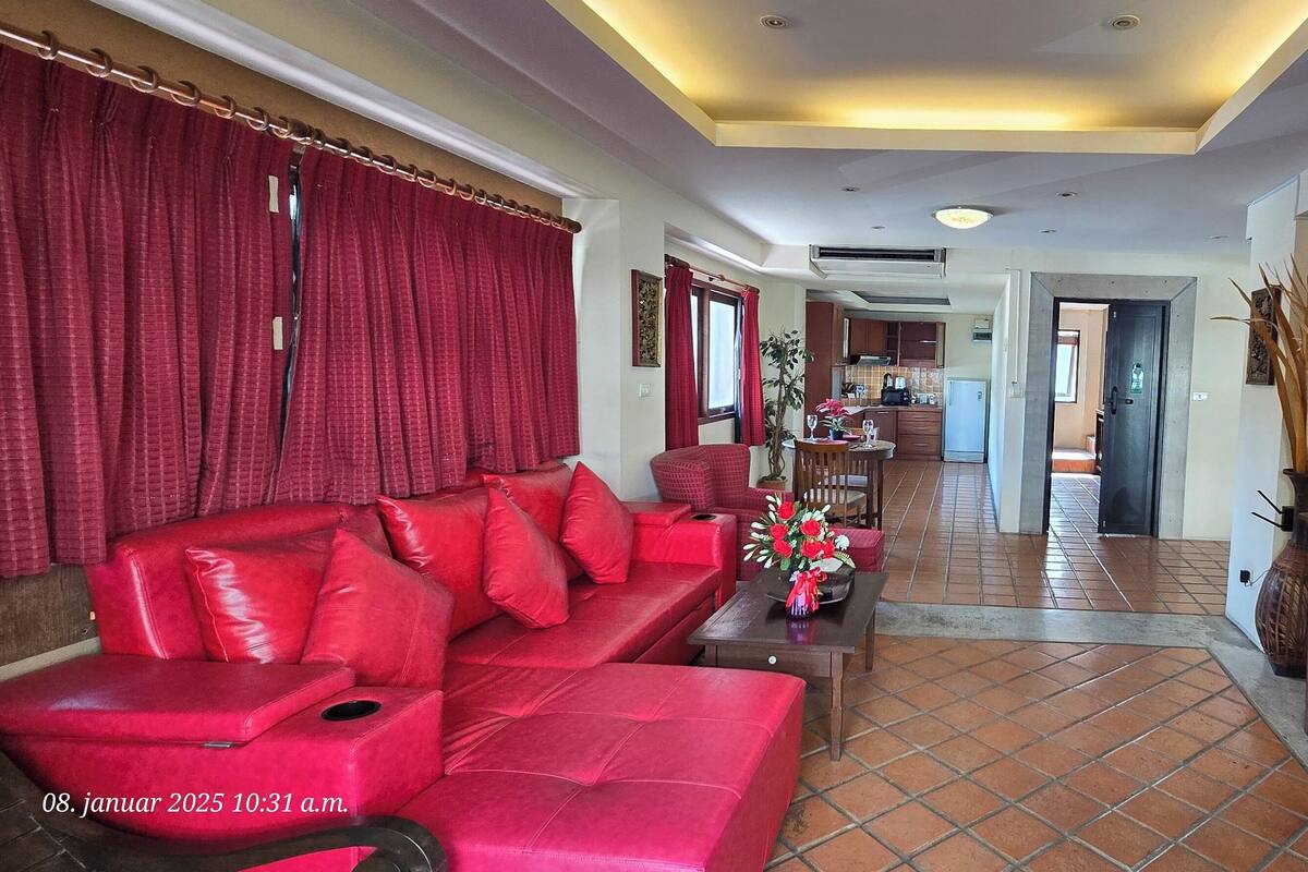 A living room with a red sofa, a coffee table with flowers, a TV on a wooden stand, and a kitchen in the background.