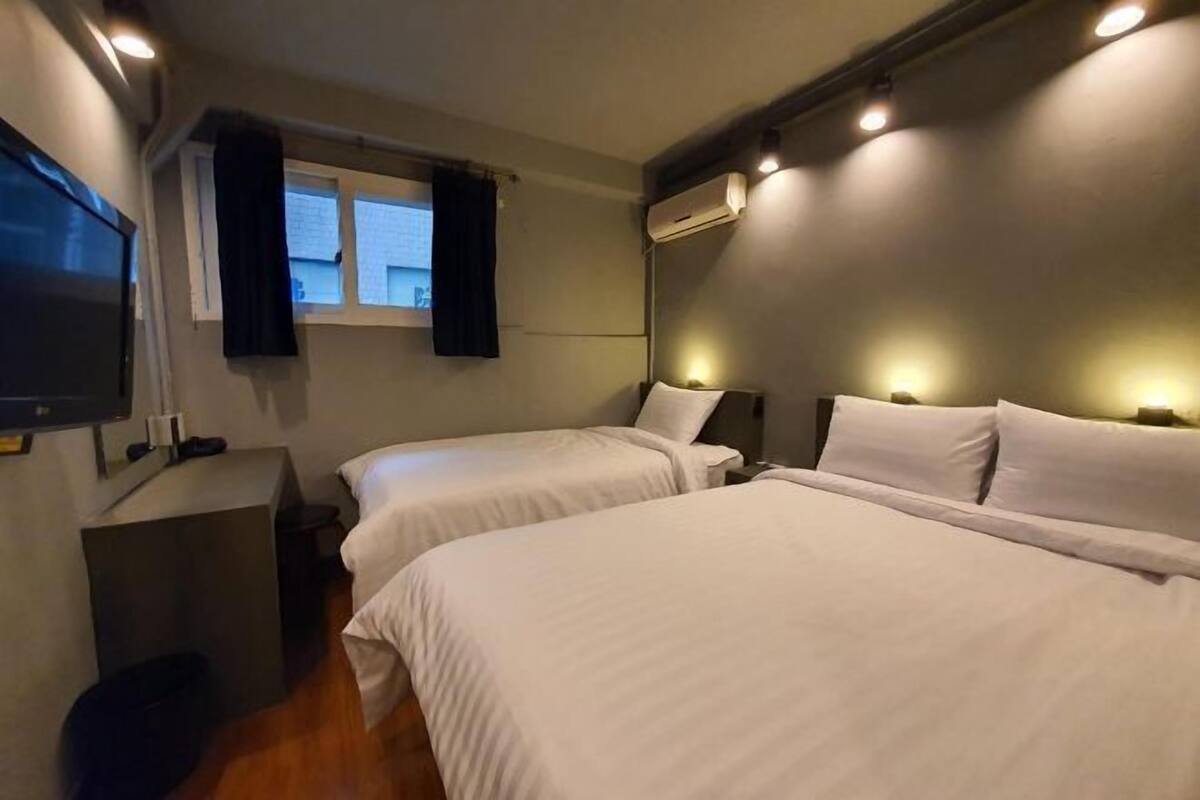 A hotel room with two beds, a TV, a window with curtains, and an air conditioning unit.
