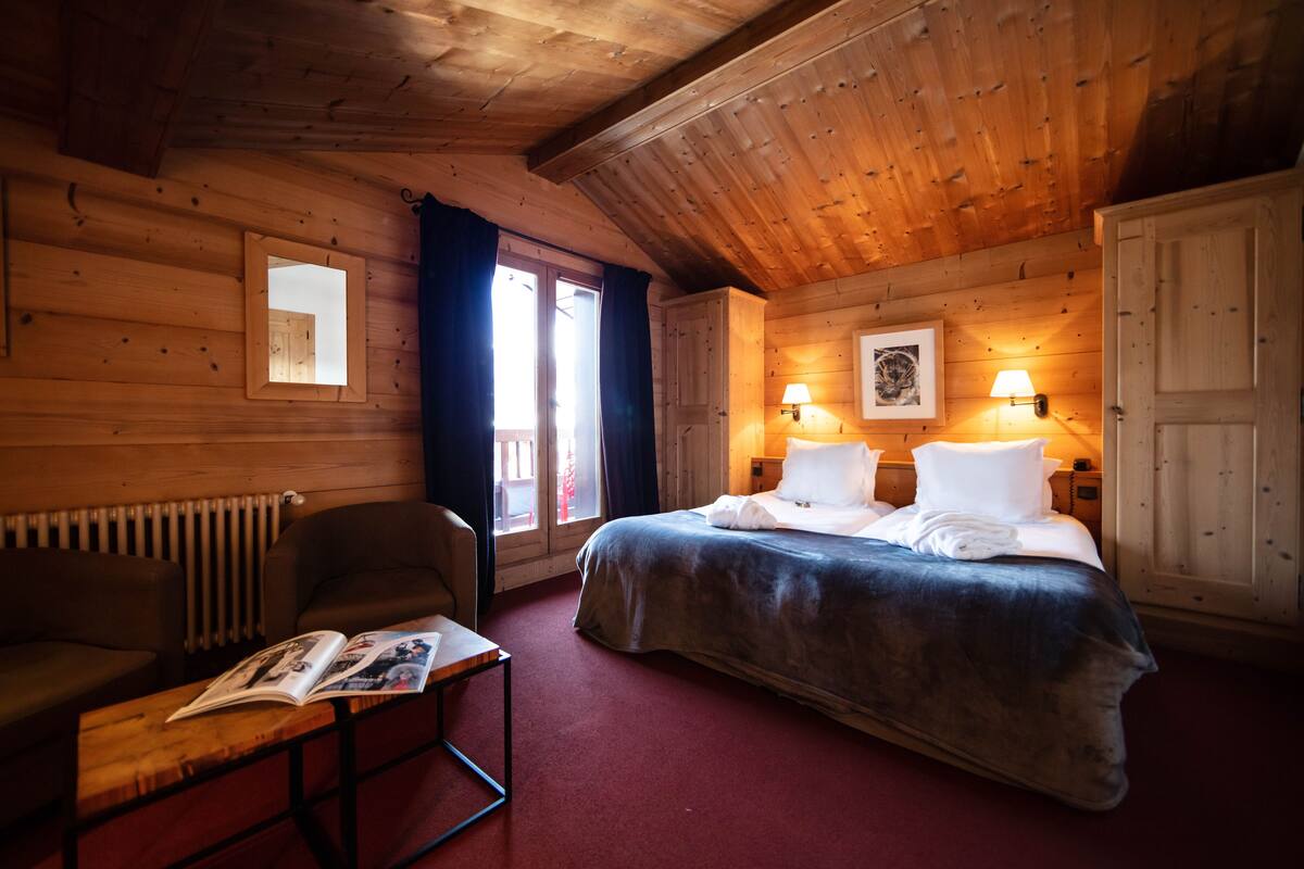 A wooden cabin room with a bed, a chair, a table with a magazine, and two wall-mounted lamps.