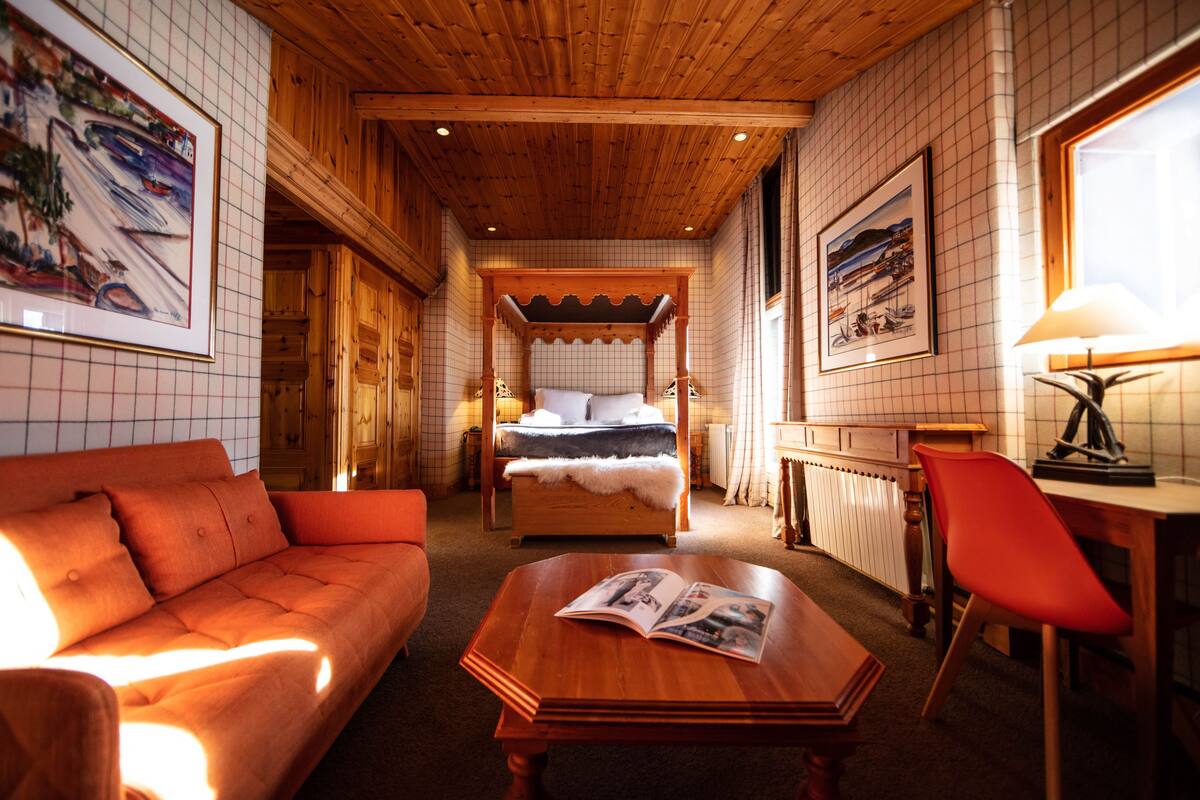A cozy room with a wooden ceiling, a bed with a white canopy, a sofa, a table with a magazine, and a chair.