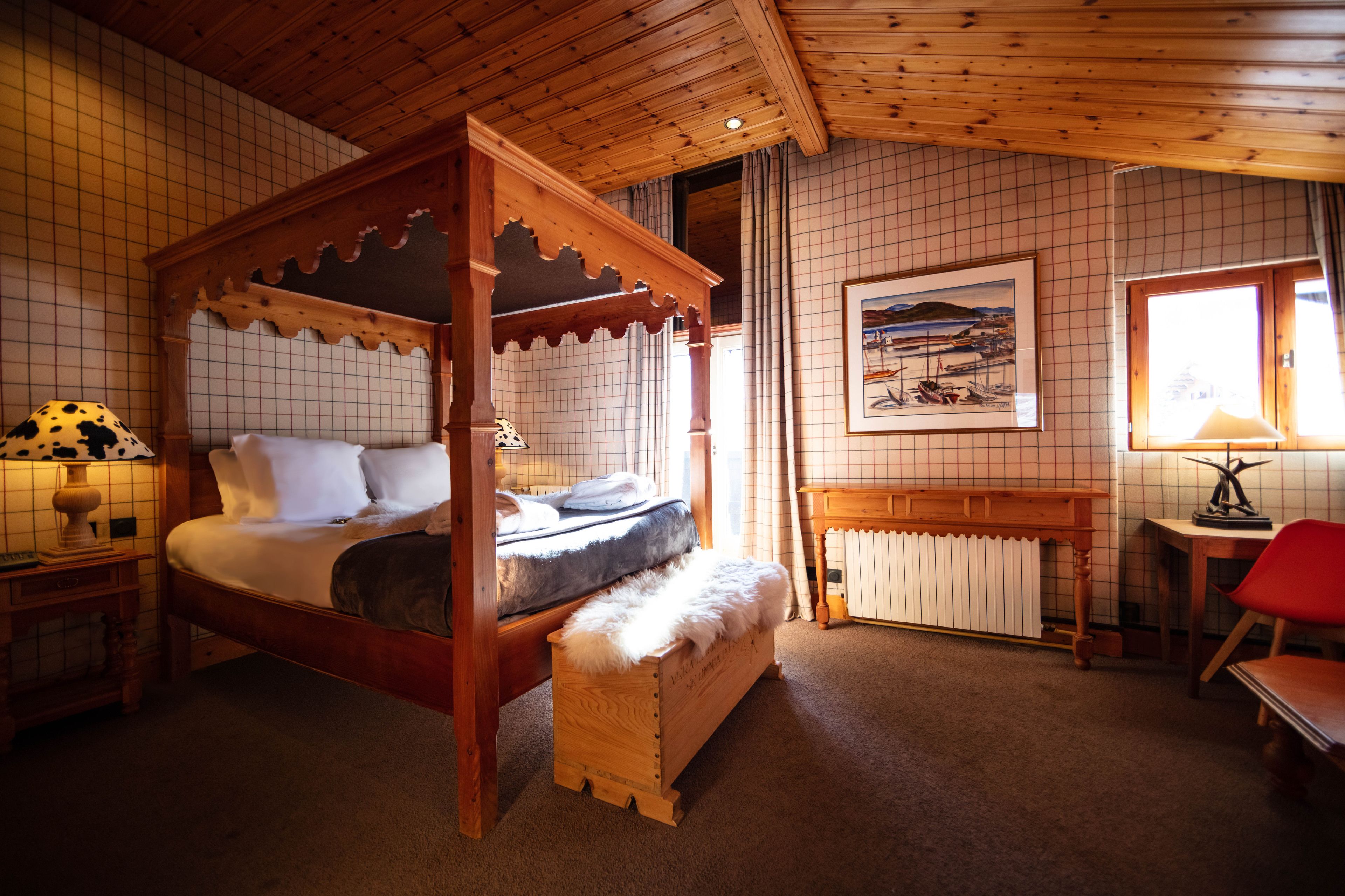 A four-poster bed with a wooden headboard, a bedside table with a lamp, a red chair, and a framed picture on the wall.