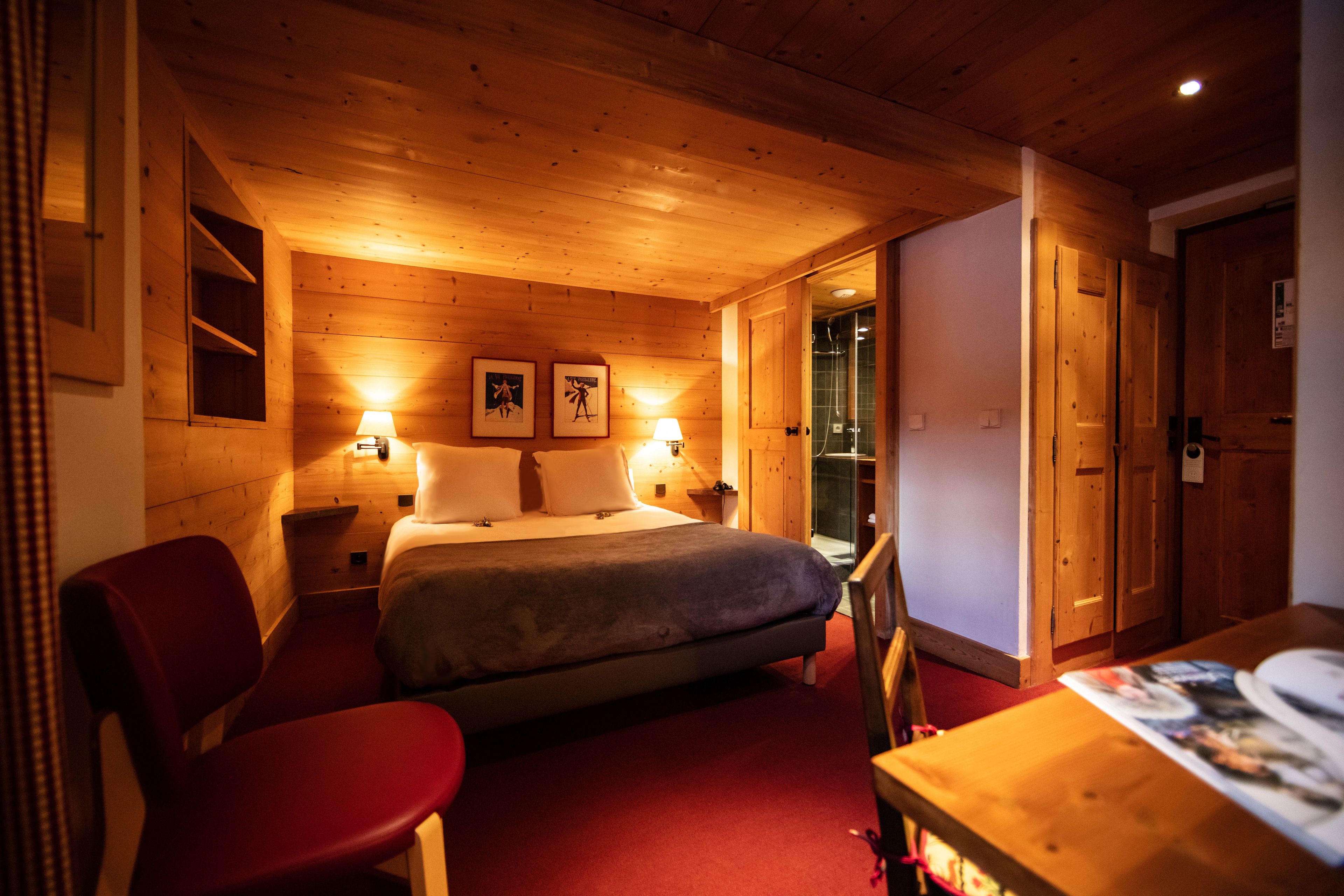 A wooden room with a bed, two bedside lamps, a red chair, and a table with a magazine.