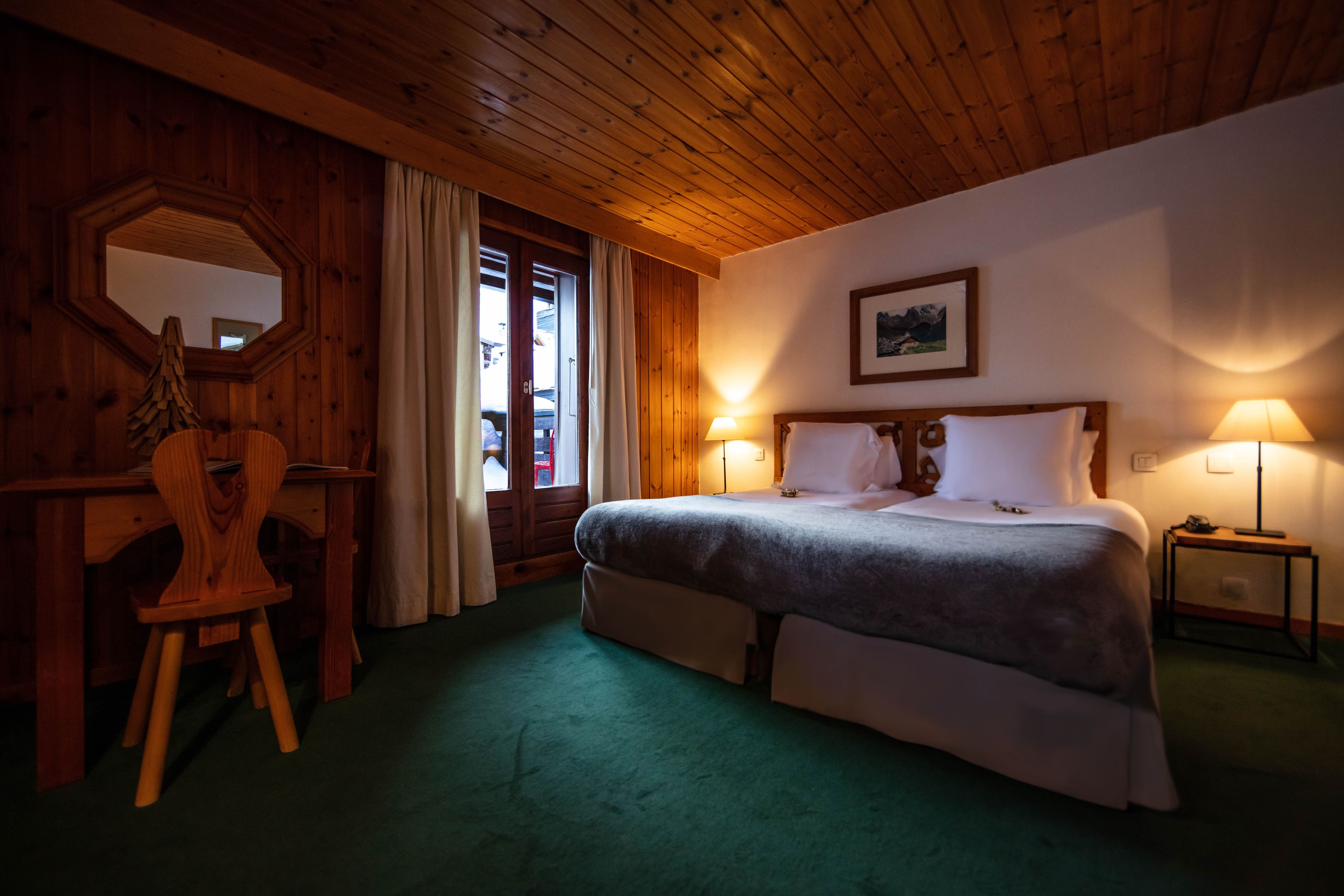 A cozy room with a wooden ceiling, a bed with white bedding, two bedside lamps, a wooden desk with a chair, and a large mirror on the wall.