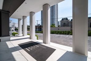 A modern hotel entrance with a black doormat, white columns, and a view of a parking lot and buildings in the background.