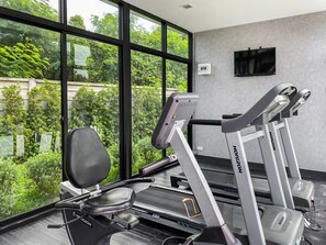 A gym with treadmills and weight machines, large windows with a view of greenery, and a flat-screen TV on the wall.