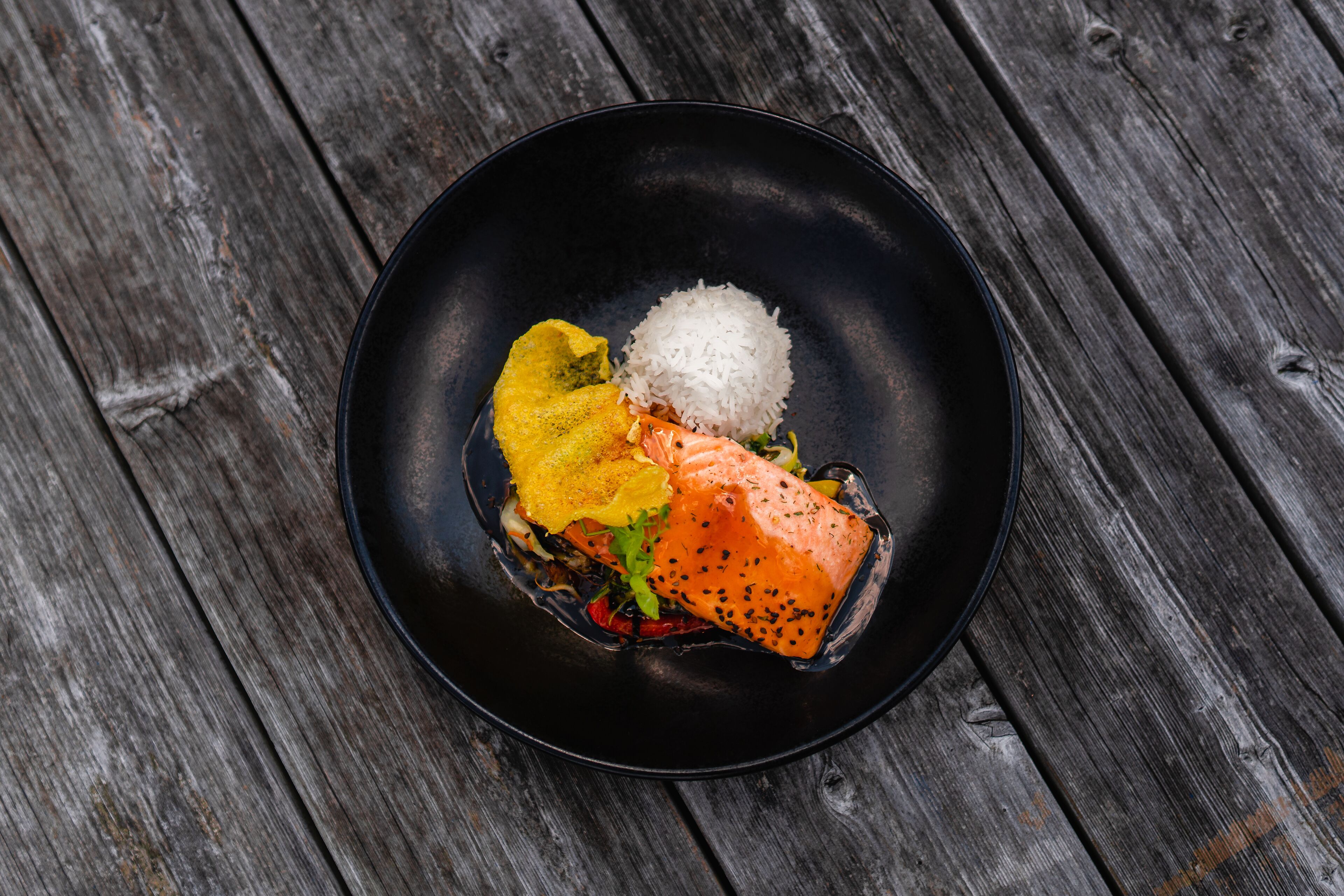 A black plate with white rice, a piece of salmon, and a yellow vegetable on a wooden surface.