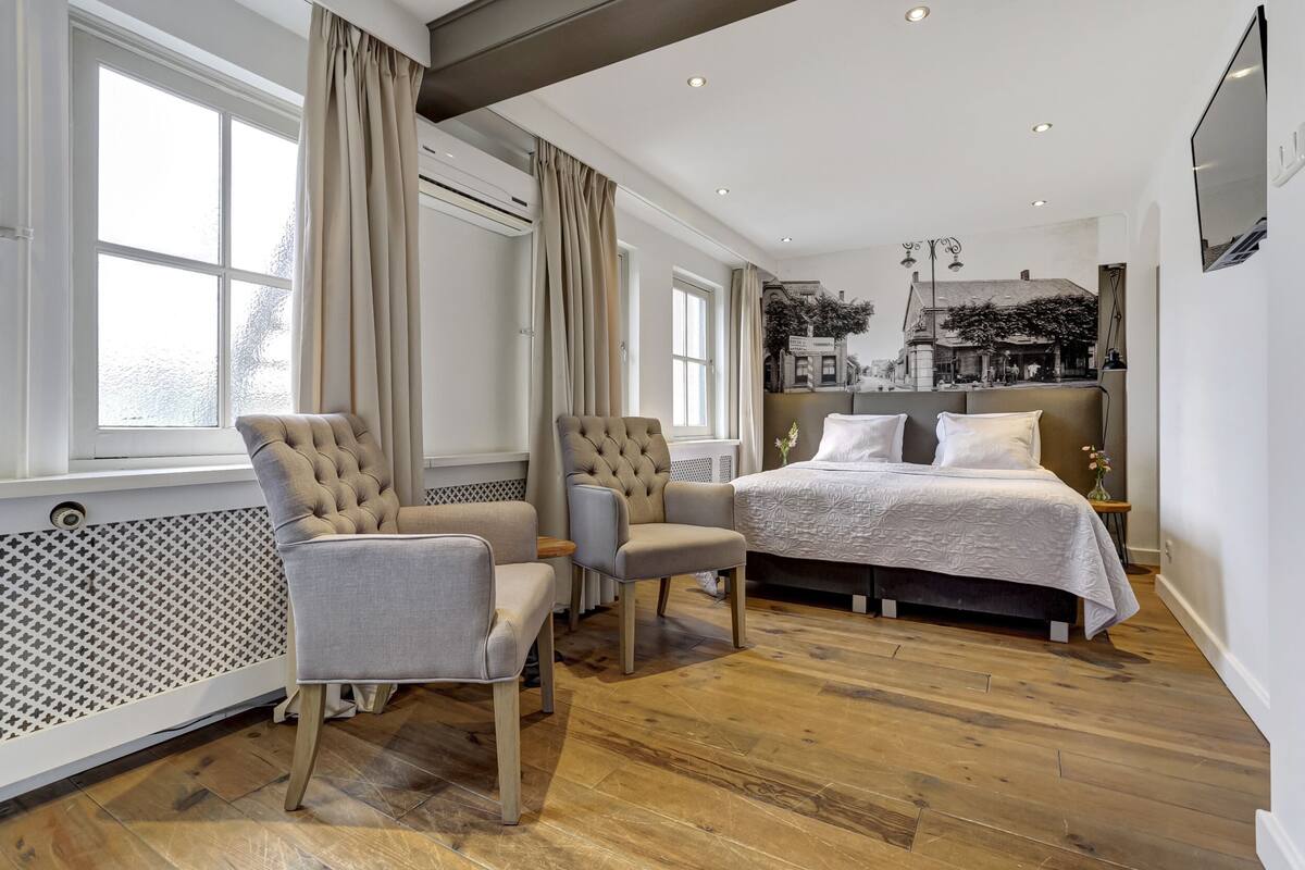 A modern bedroom with a large bed, two armchairs, a radiator, and a view of a cityscape through the window.