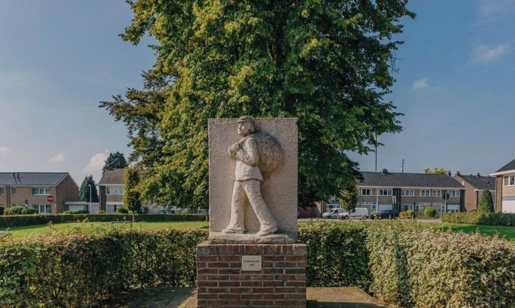 A statue of a person carrying a large object, set in a landscaped area with a hedge and a tree in the background.