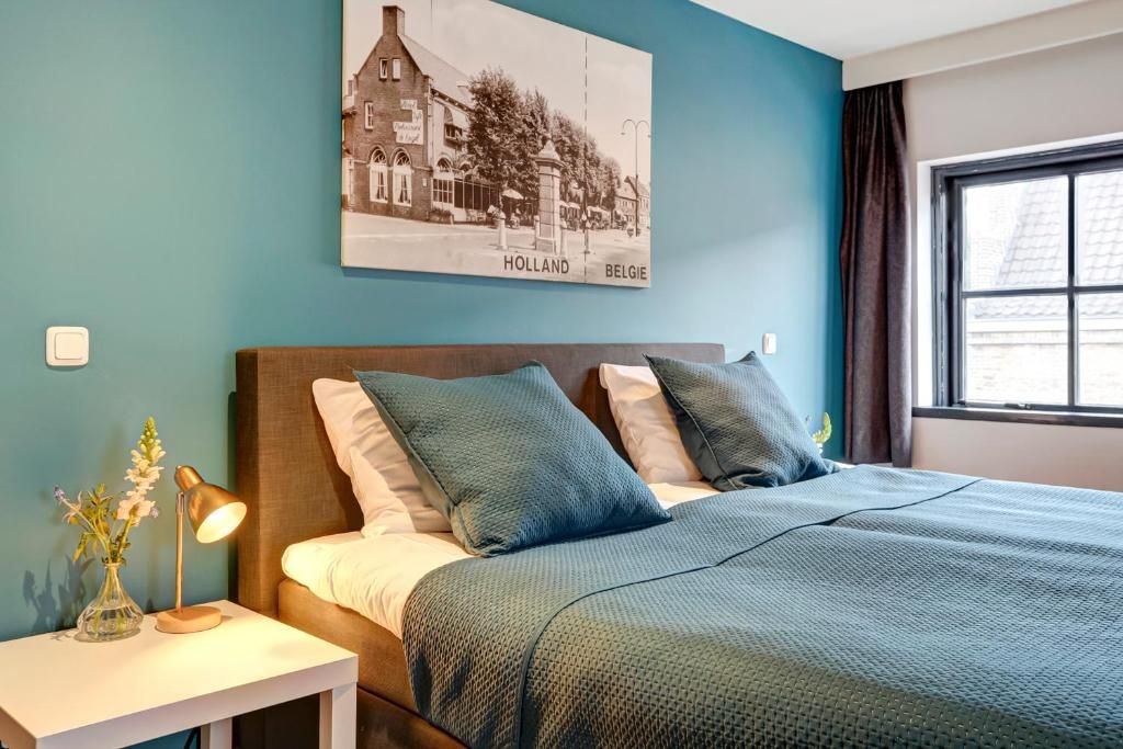 Un dormitorio con una cama, almohadas, una mesita de noche con una lámpara y un cuadro colgado en la pared.