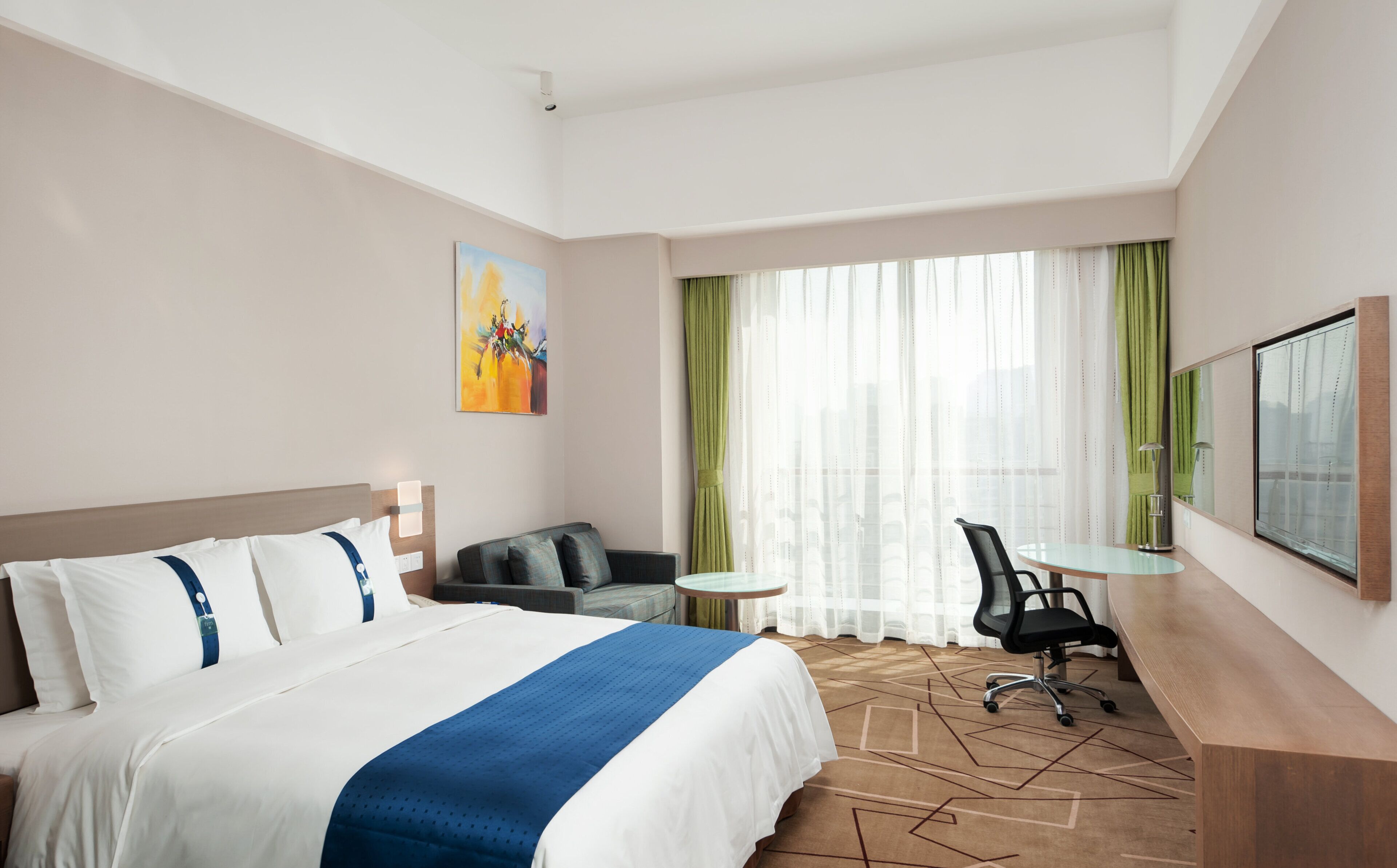 A hotel room with a bed, a desk, a chair, a small table, and a large window with curtains.
