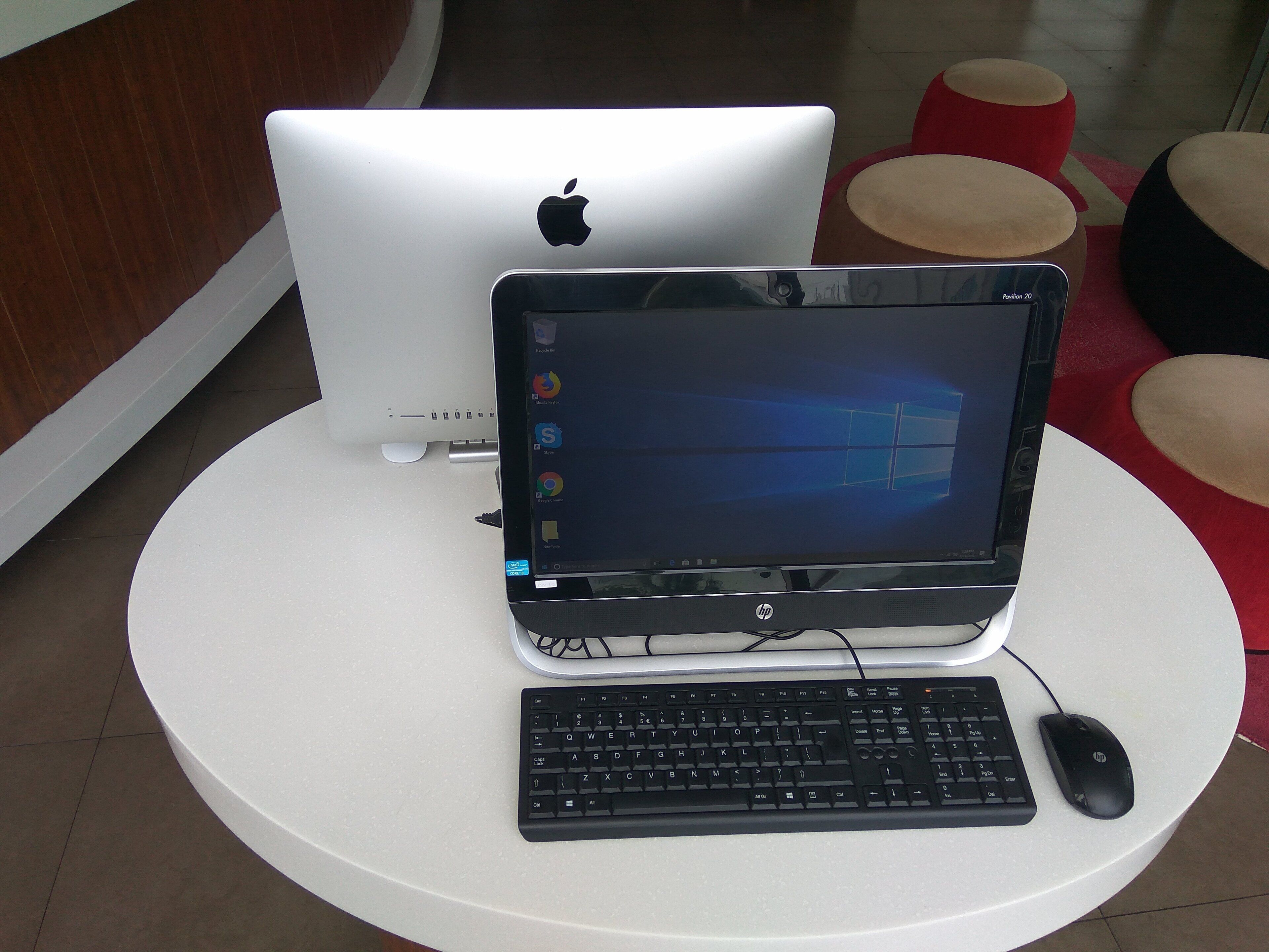 Un portatile su un tavolo bianco, con tastiera e mouse, un computer desktop Apple bianco e pouf rossi e neri sullo sfondo.