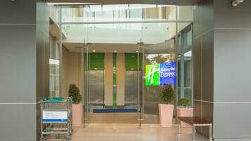 A hotel entrance with a glass door, a digital signboard, a bench, and a shopping cart.